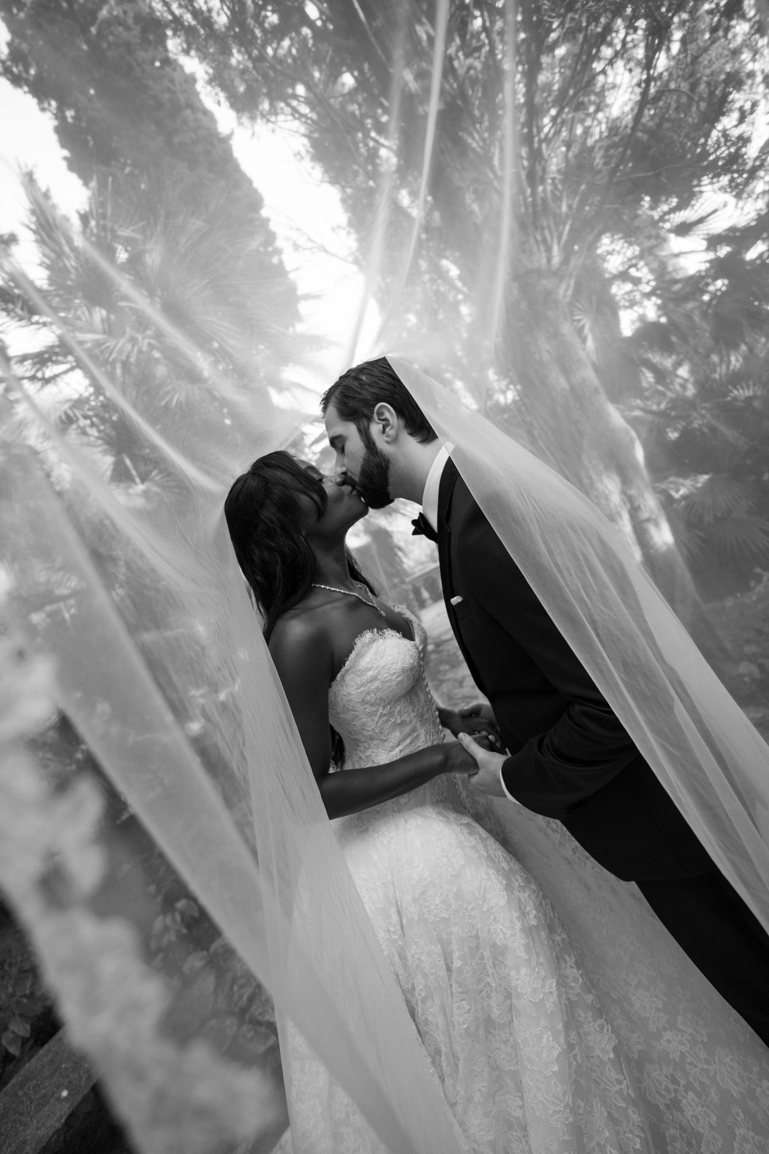 Black and white photo of a bride and groom kissing tenderly beneath a flowing sheer wedding veil outdoors.