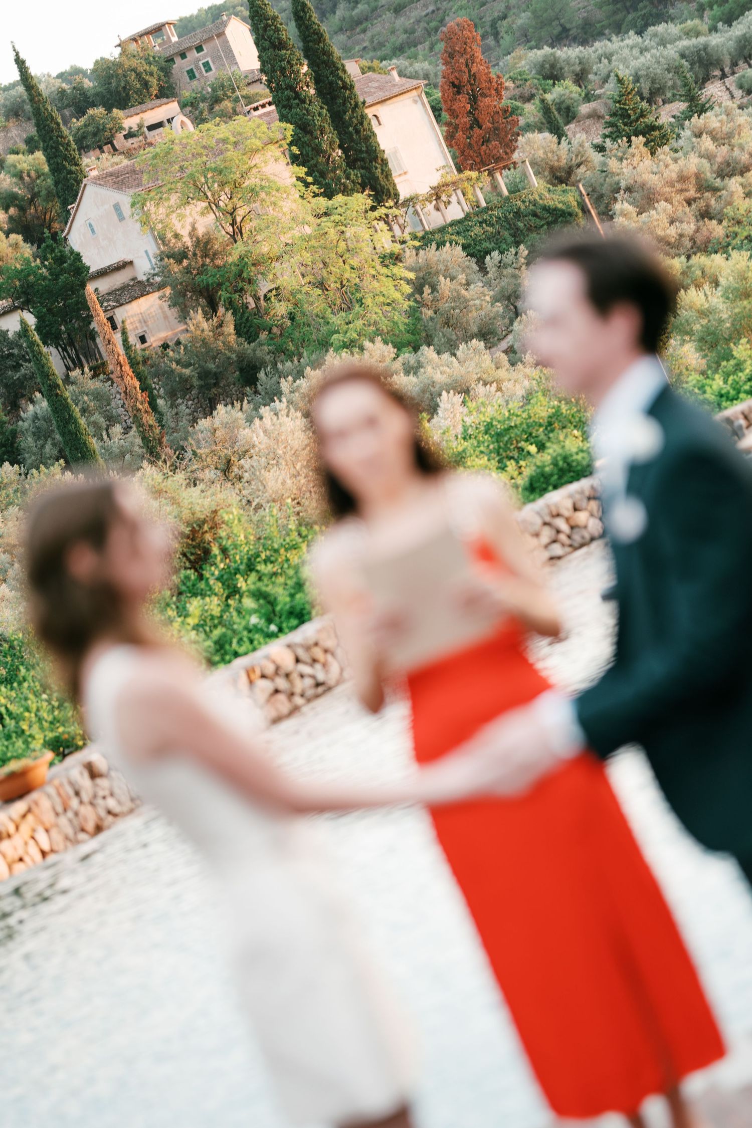 Blurred wedding guests in a red dress and dark suit stand on a stone terrace overlooking a lush Mediterranean hillside village.