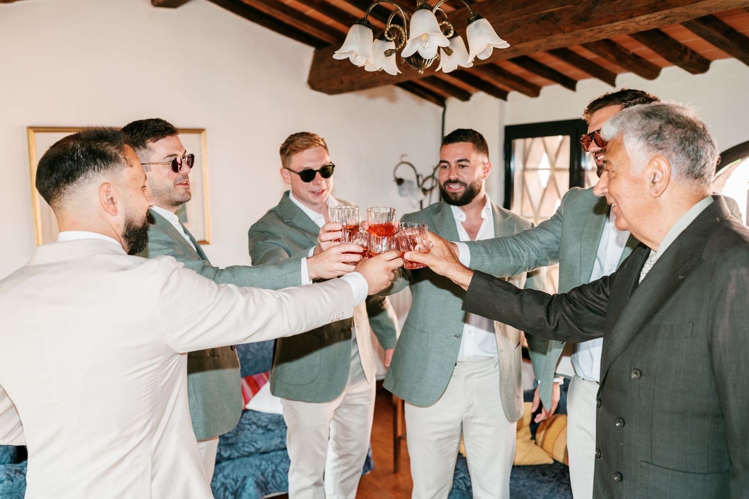 Group of men in suits and sunglasses raising glasses of red drinks in a cheerful toast inside a rustic room with wooden beams.