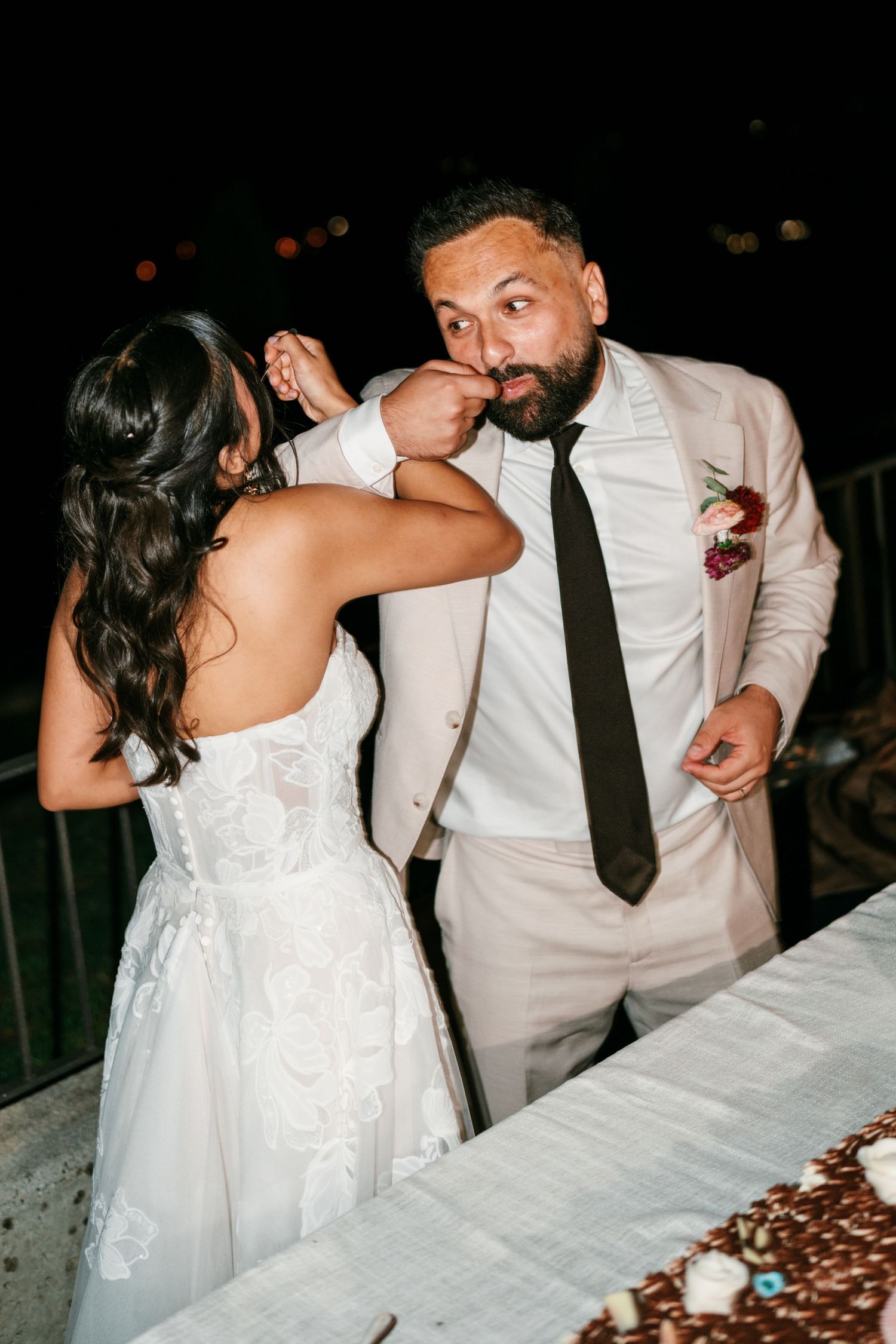 A bride in a white lace gown playfully feeds wedding cake to a groom in a beige suit and dark tie at a nighttime outdoor reception.