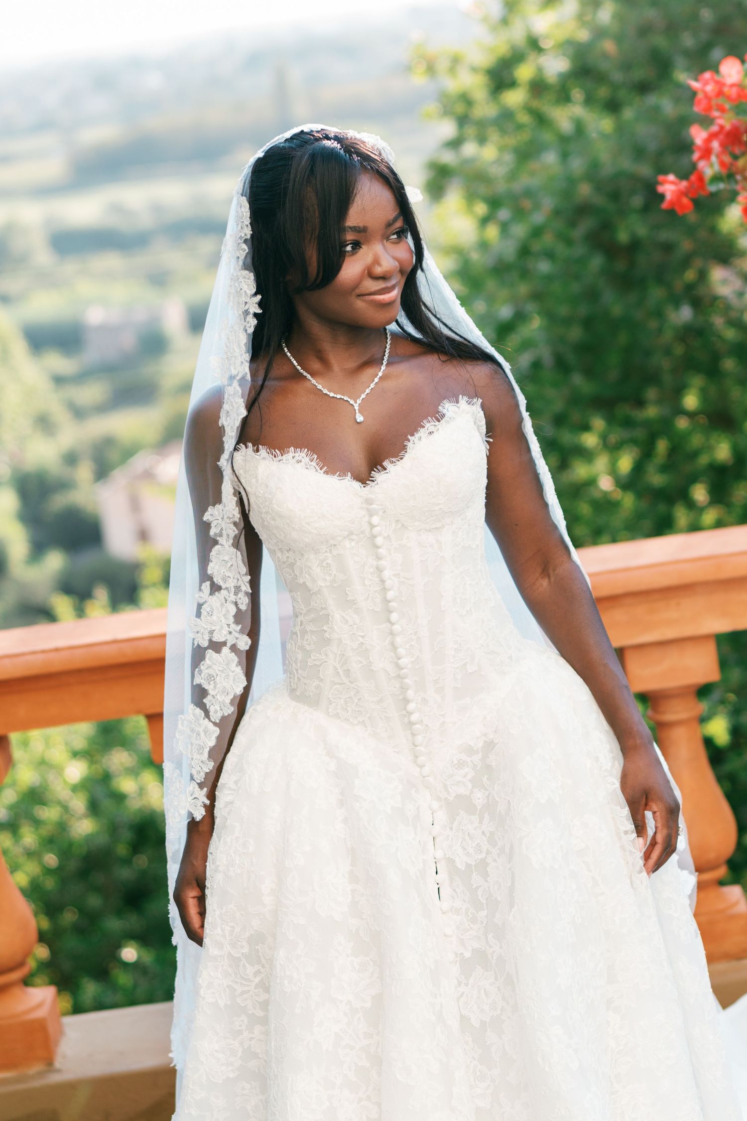 Smiling bride in a white lace sweetheart wedding gown and long veil poses on a terracotta balcony overlooking green hills.