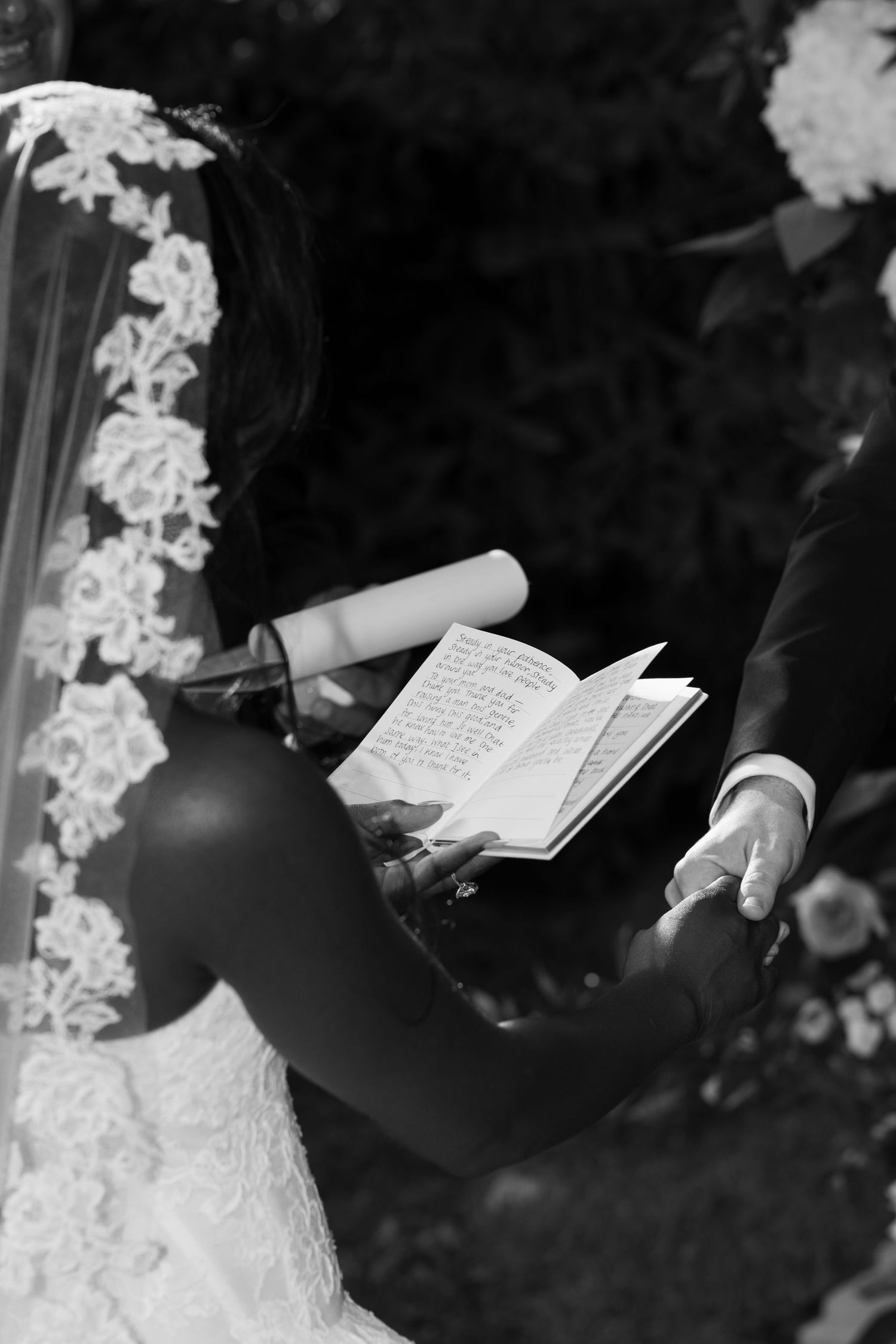 Black and white photo of a bride in a lace veil holding hands with her groom as he reads handwritten vows from an open book.