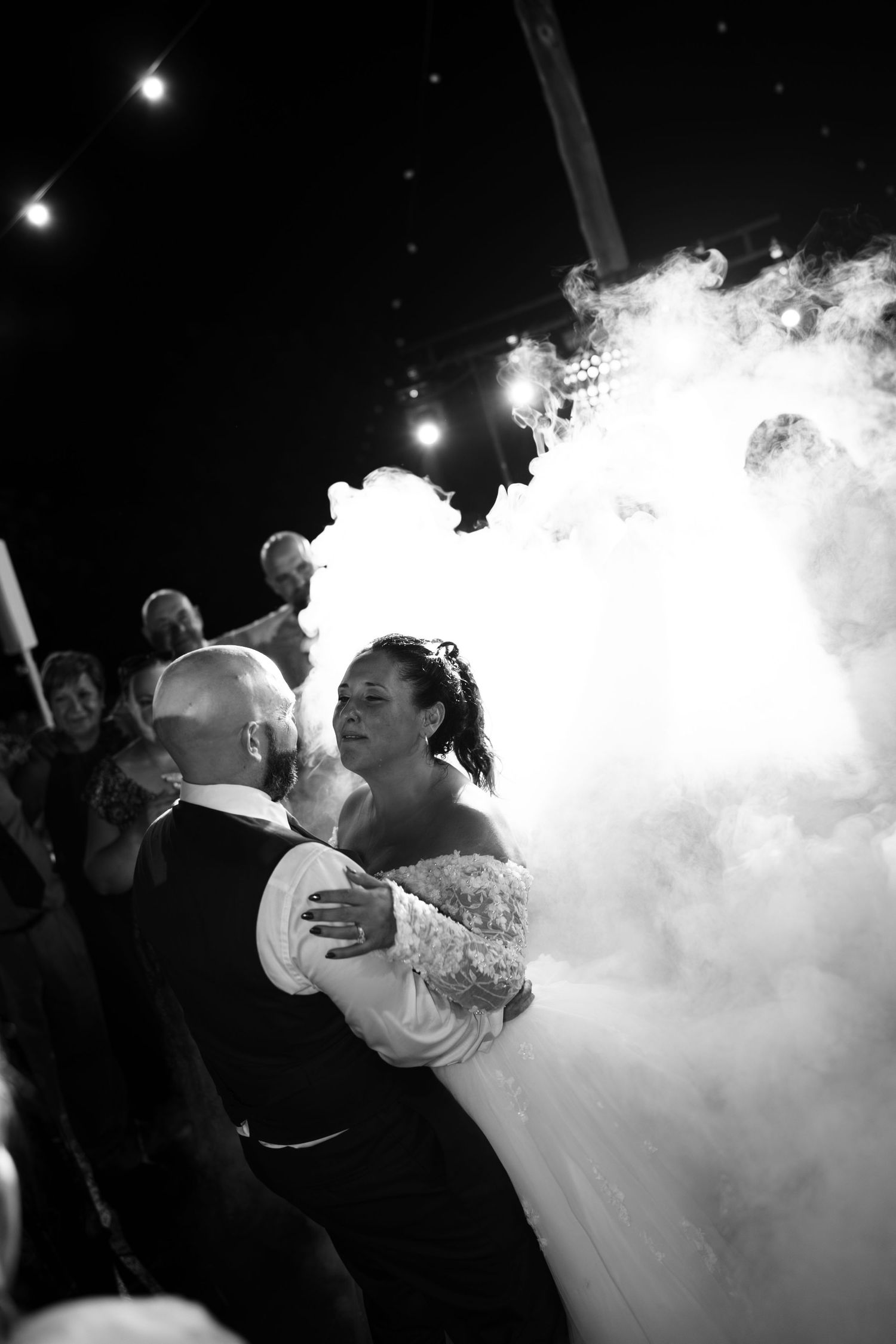 Black and white photo of a bride and groom sharing a first dance surrounded by thick dancing fog as guests watch behind them.