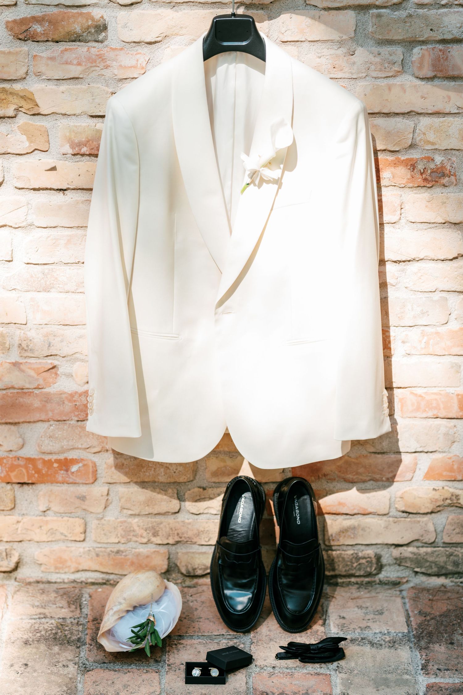 White wedding tuxedo jacket with boutonniere hanging on brick wall above black loafers, cufflinks, and accessories.
