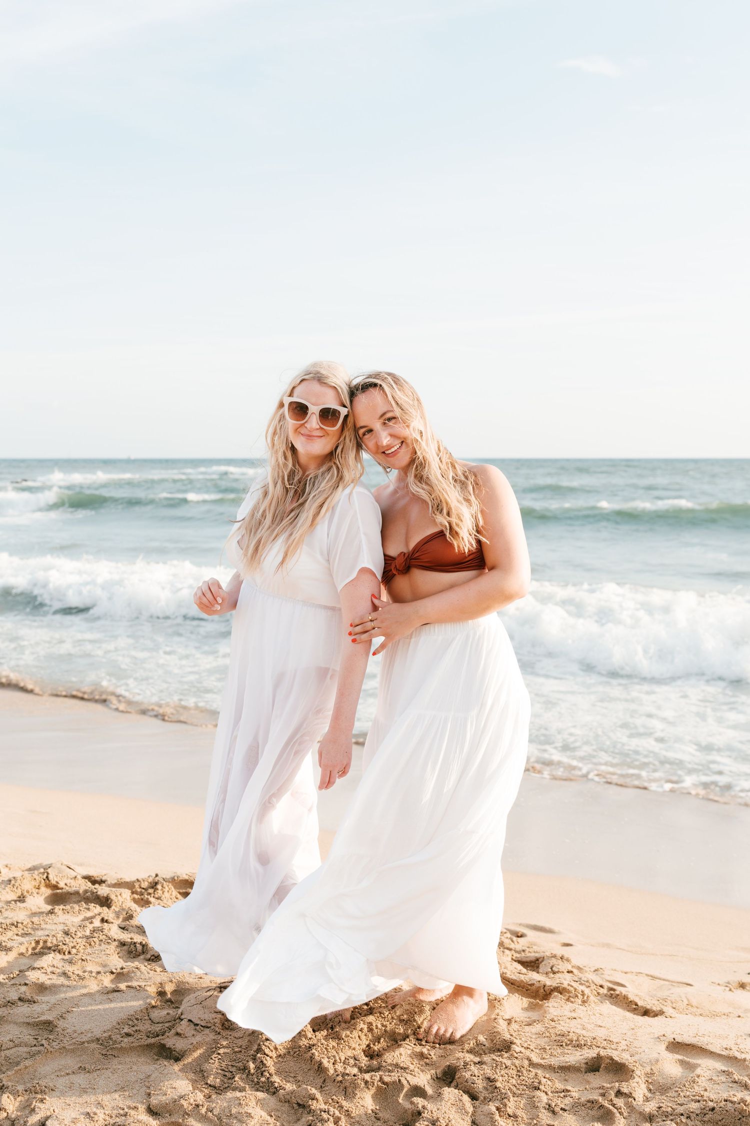 Two blonde women in white dresses embrace on a sunny beach with ocean waves behind them. Mallorca wedding filmmaker, videographer