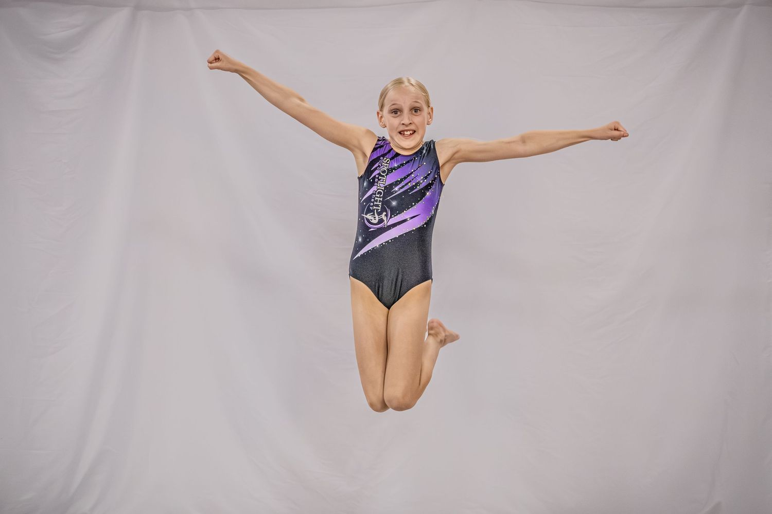 Young girl in purple gymnastics leotard jumps mid-air with arms spread wide.