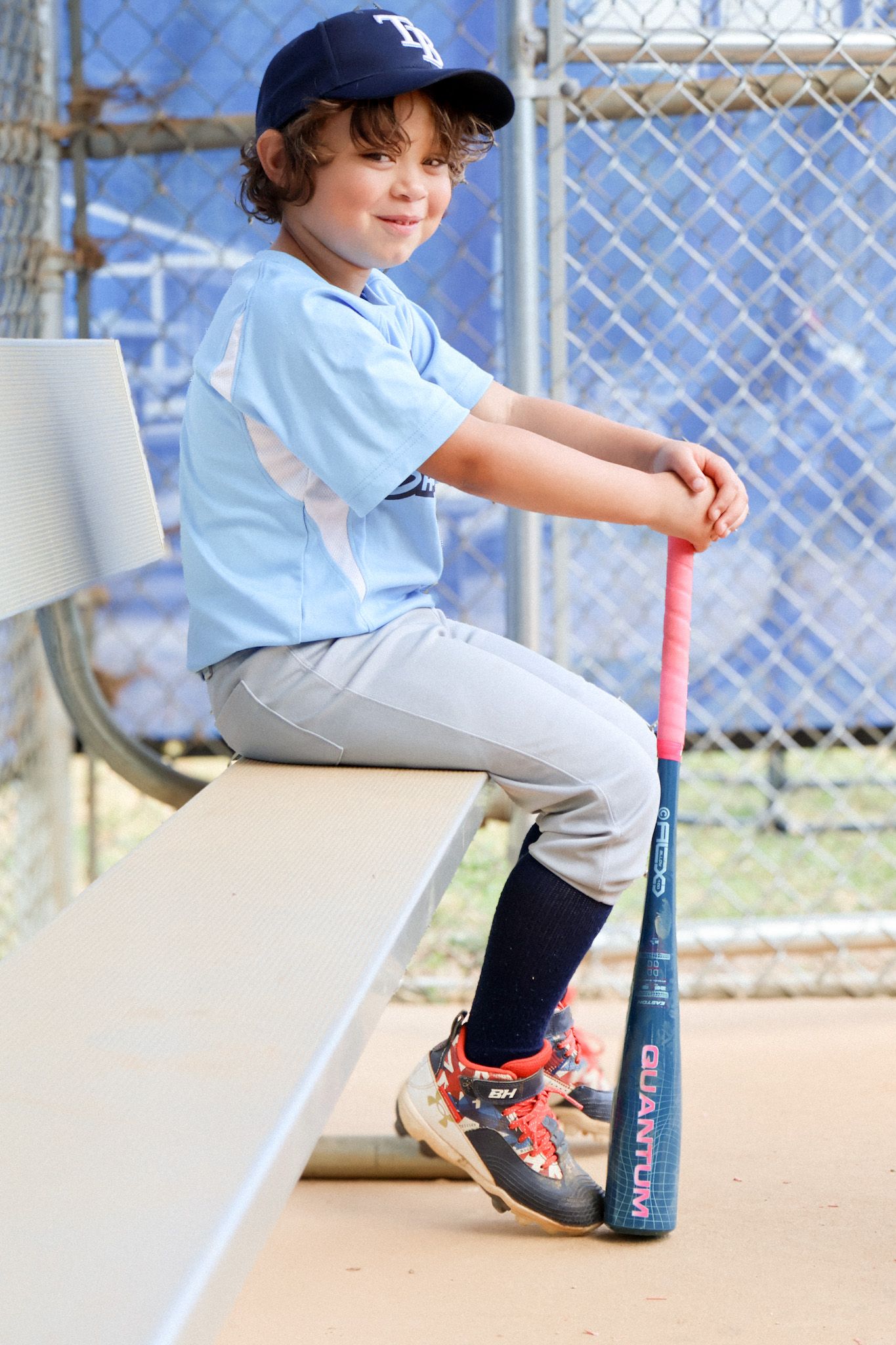 Young baseball player in Tampa Bay uniform sits on dugout bench holding bat.