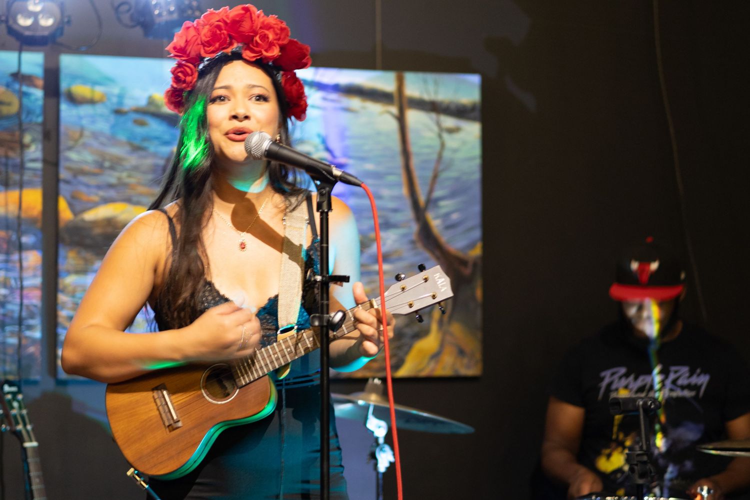 Female musician with red flower crown plays ukulele and sings at indoor venue.
