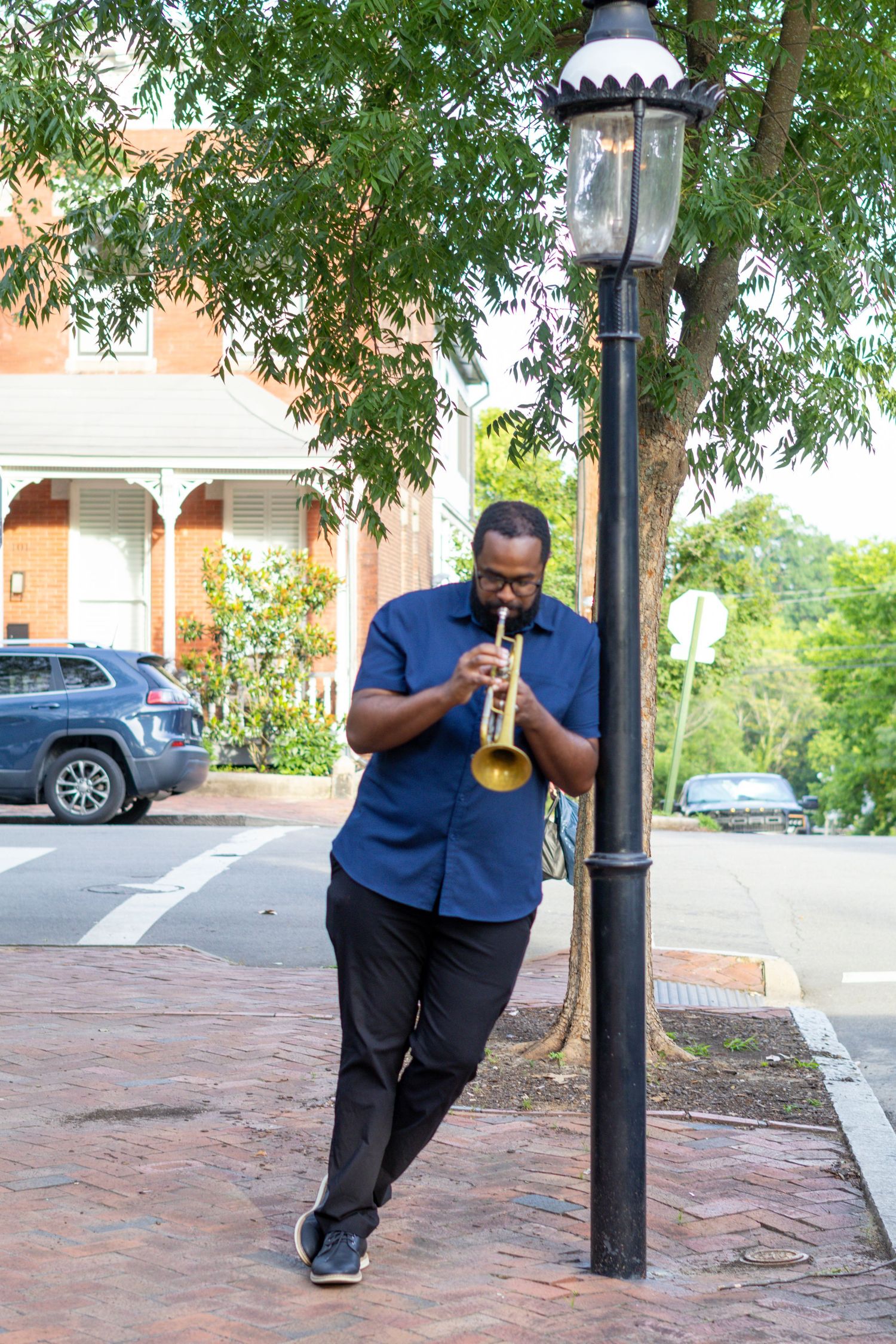 A Black man in a blue shirt plays trumpet while leaning against a street lamp on a brick sidewalk.