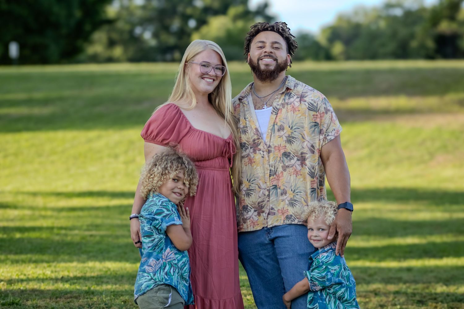 Smiling family of four poses together on lush green lawn in outdoor park.