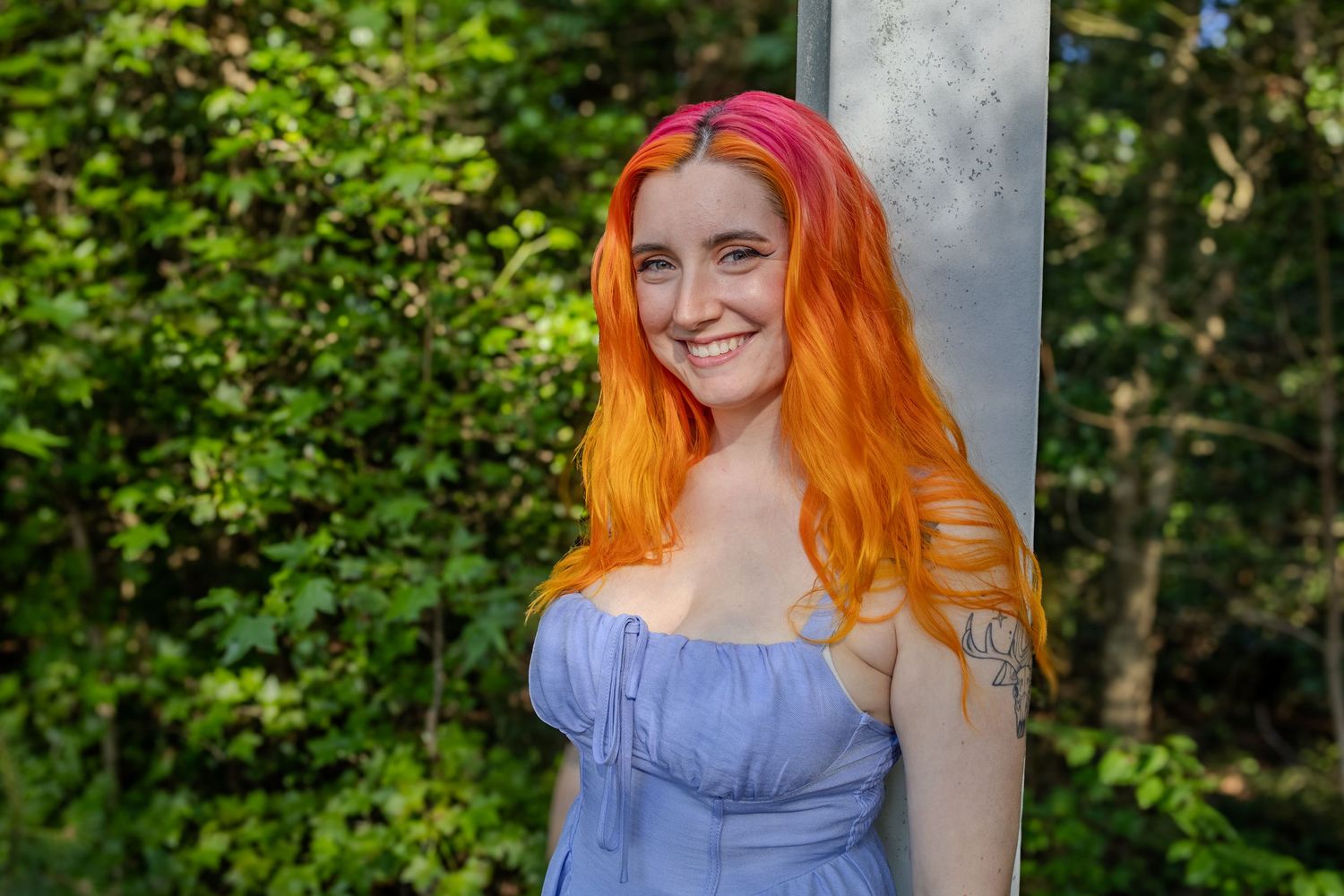 A smiling young woman with vibrant orange and pink hair leans against a metal post outdoors.