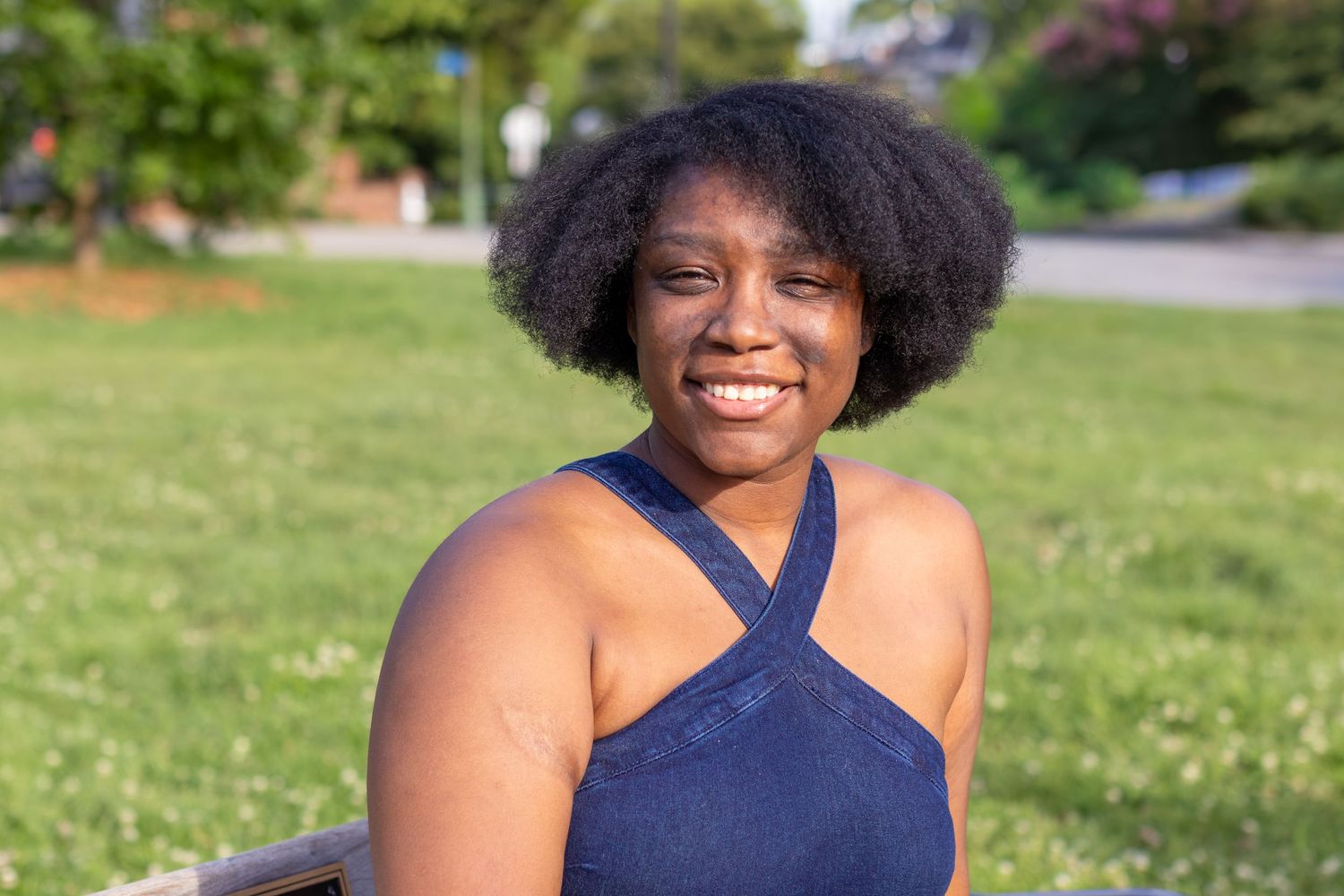 Smiling young Black woman with natural afro hair wearing a blue halter top, sitting outdoors in a sunny green park.