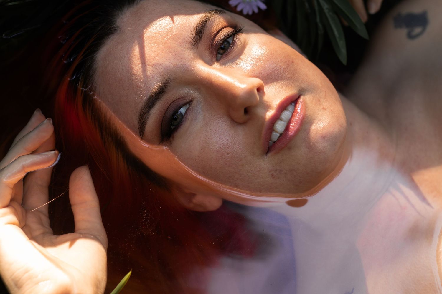 Close-up portrait of a young woman with red hair and freckles lying down, surrounded by soft natural light and flowers.