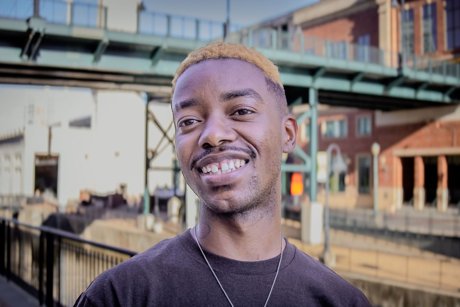 Young Black man with blonde hair smiles broadly near urban bridge overpass.
