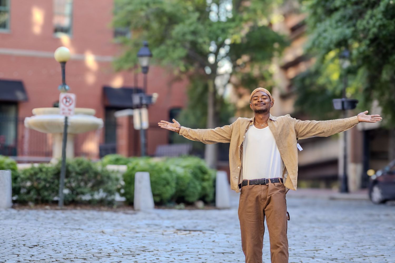Happy man in beige blazer stands with arms outstretched on cobblestone city street.