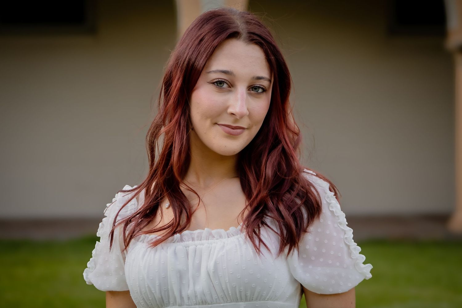 Young woman with dark red hair smiling in white off-shoulder dress outdoors.