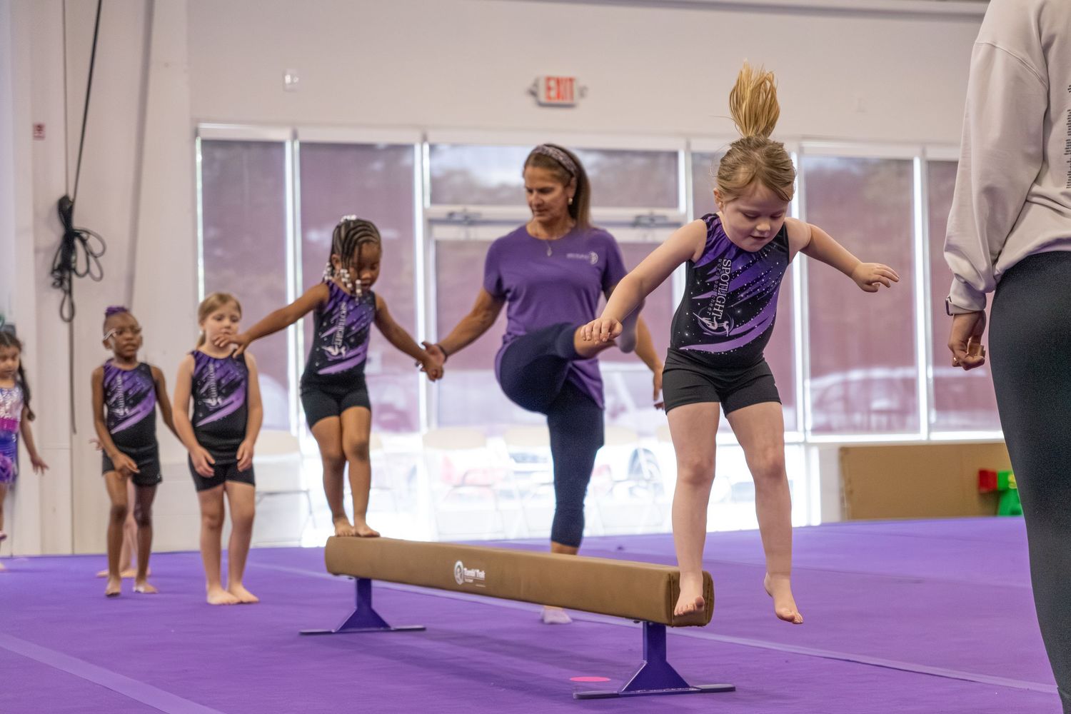 A gymnastics instructor helps a young girl balance on a low beam while other children watch.