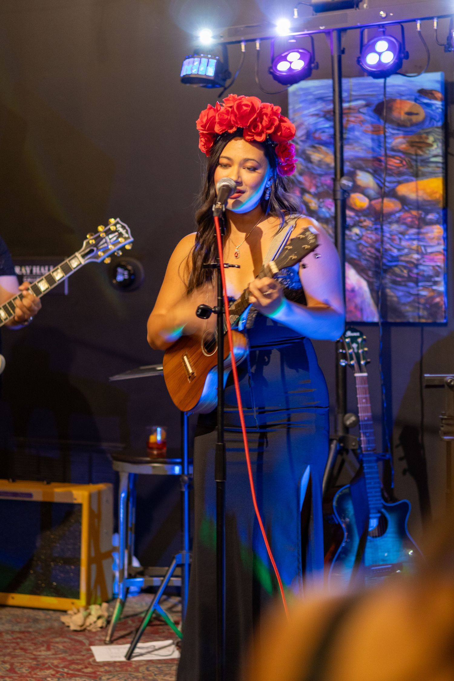 Woman with red floral crown plays ukulele and sings into microphone at live show.
