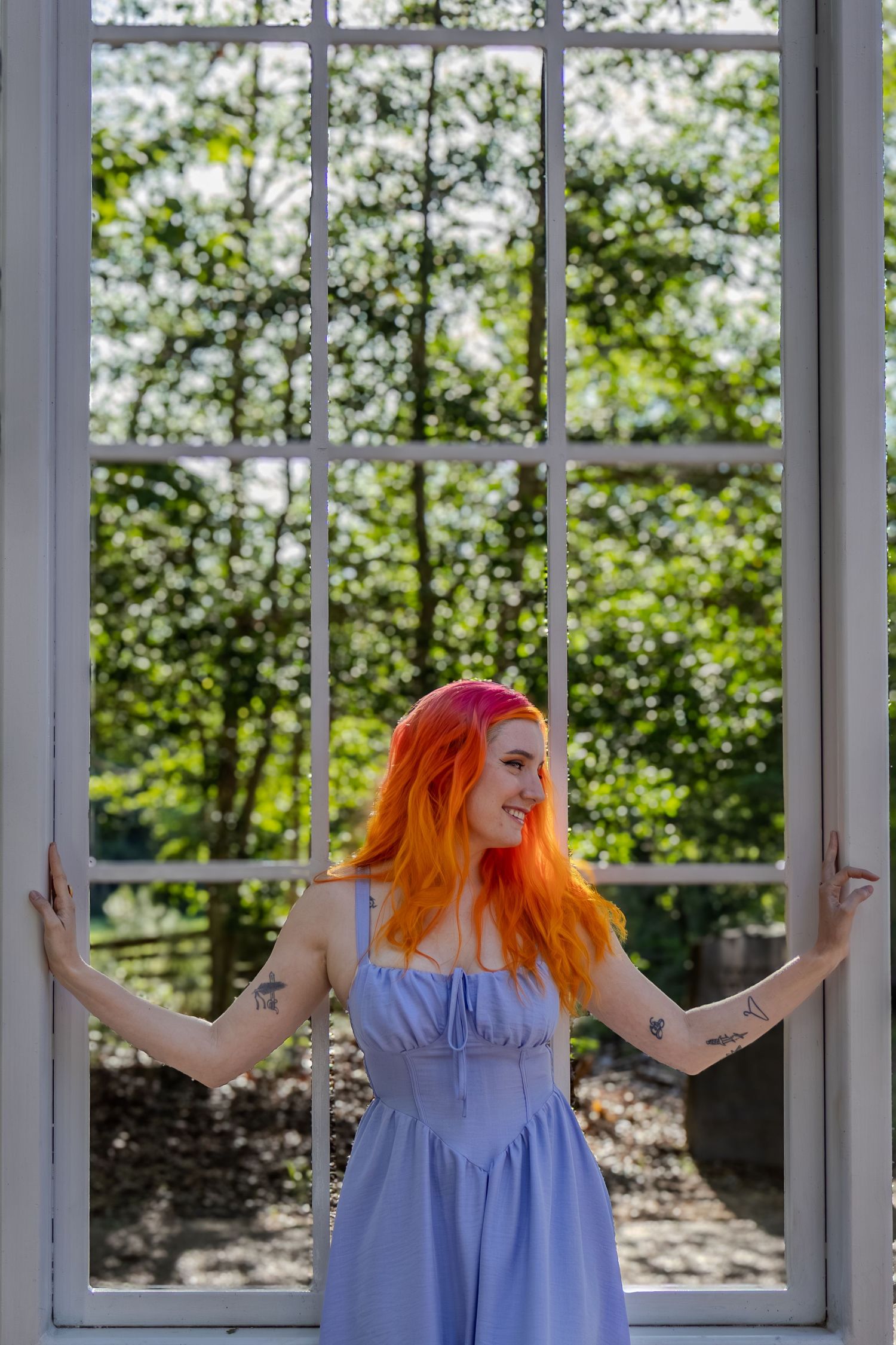 Young woman with vibrant orange-pink hair stands in open white window frame outdoors.