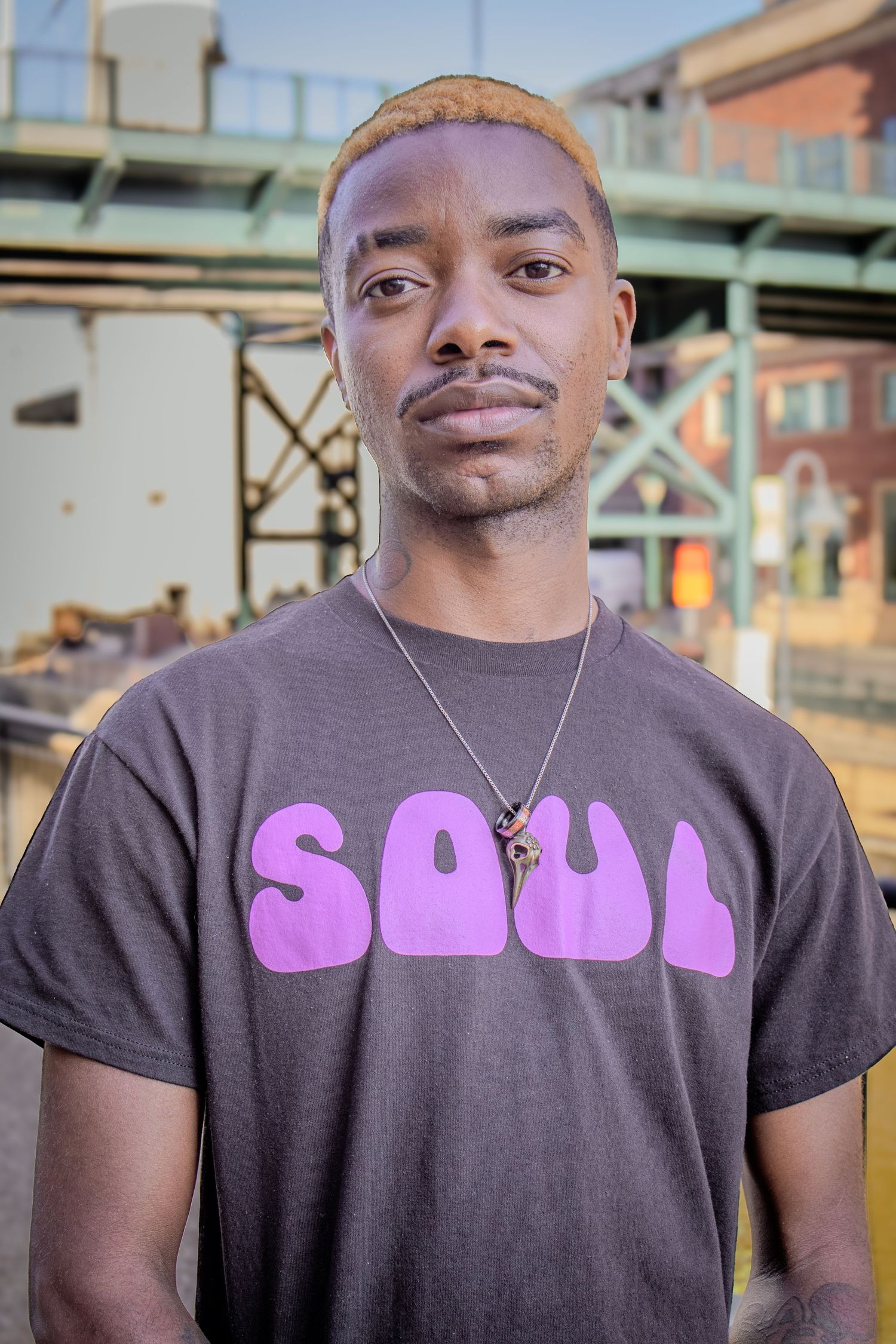 Young Black man in purple SOUL graphic tee poses outdoors near urban bridge.