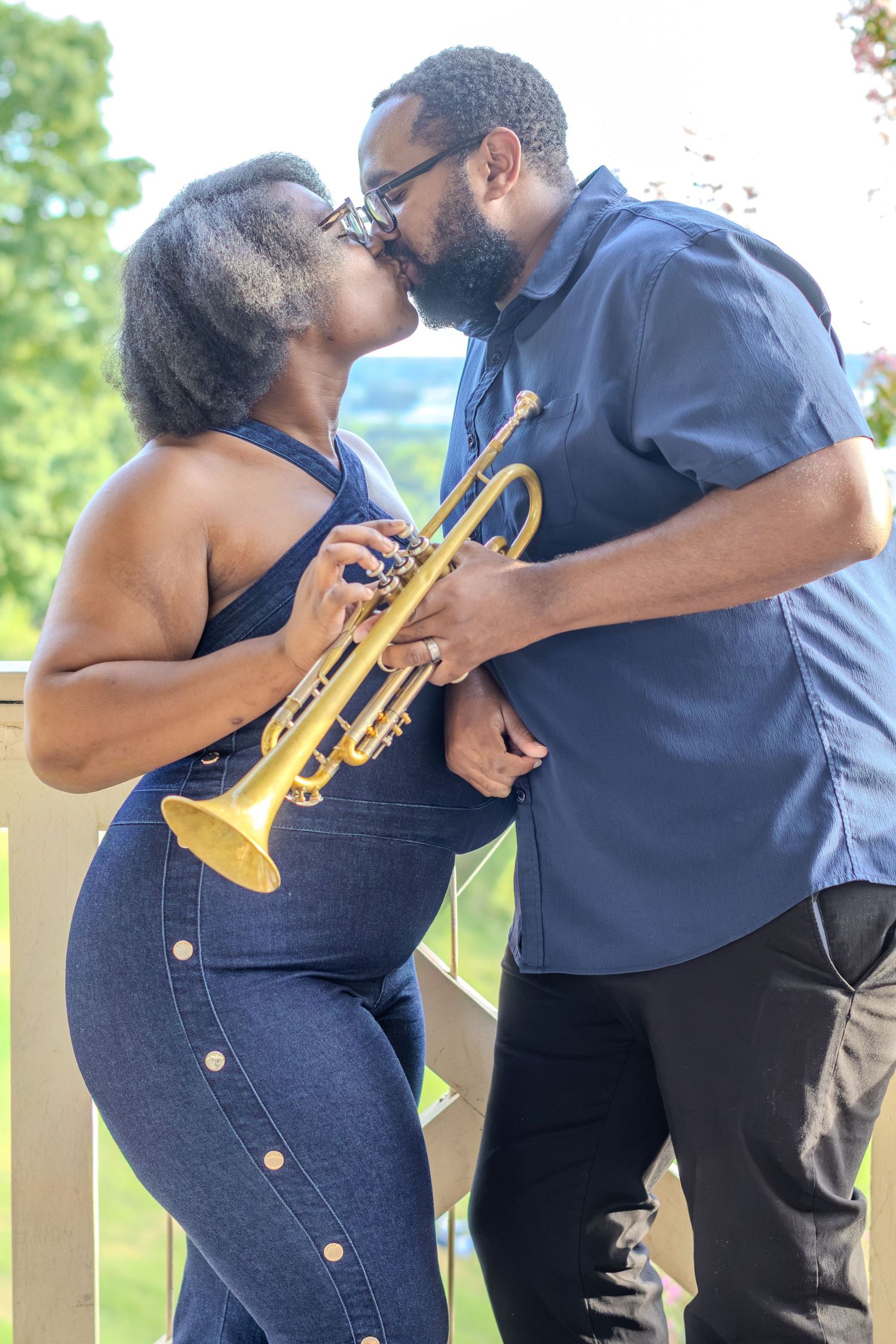 A couple shares a romantic kiss while holding a golden trumpet outdoors on a deck surrounded by lush greenery.