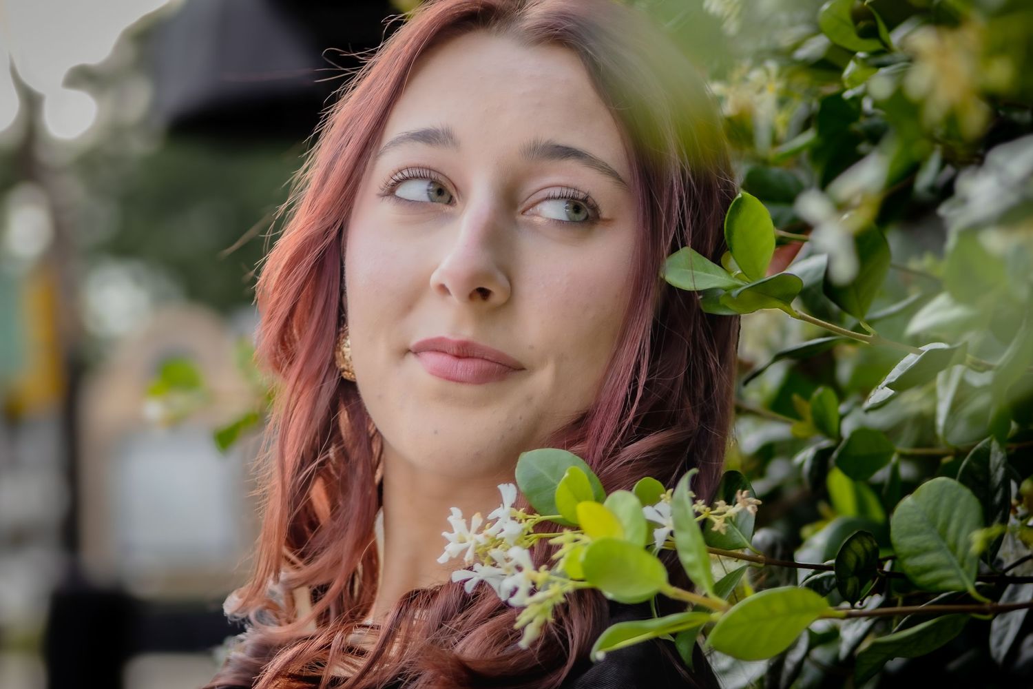 Young woman with pink hair peers through lush green flowering bushes outdoors.