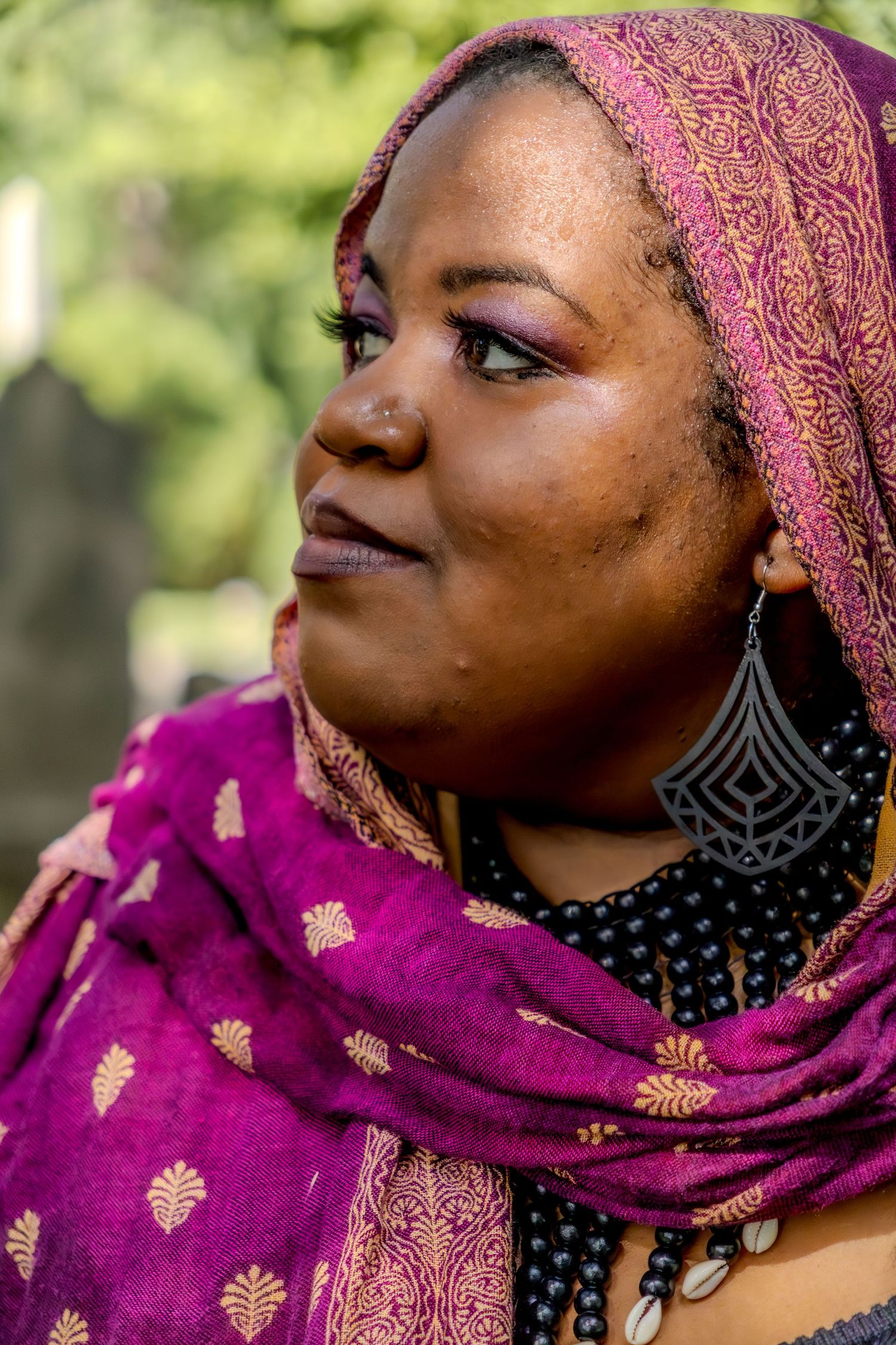 A woman wearing a purple and gold headscarf with matching earrings and makeup gazes thoughtfully outdoors.