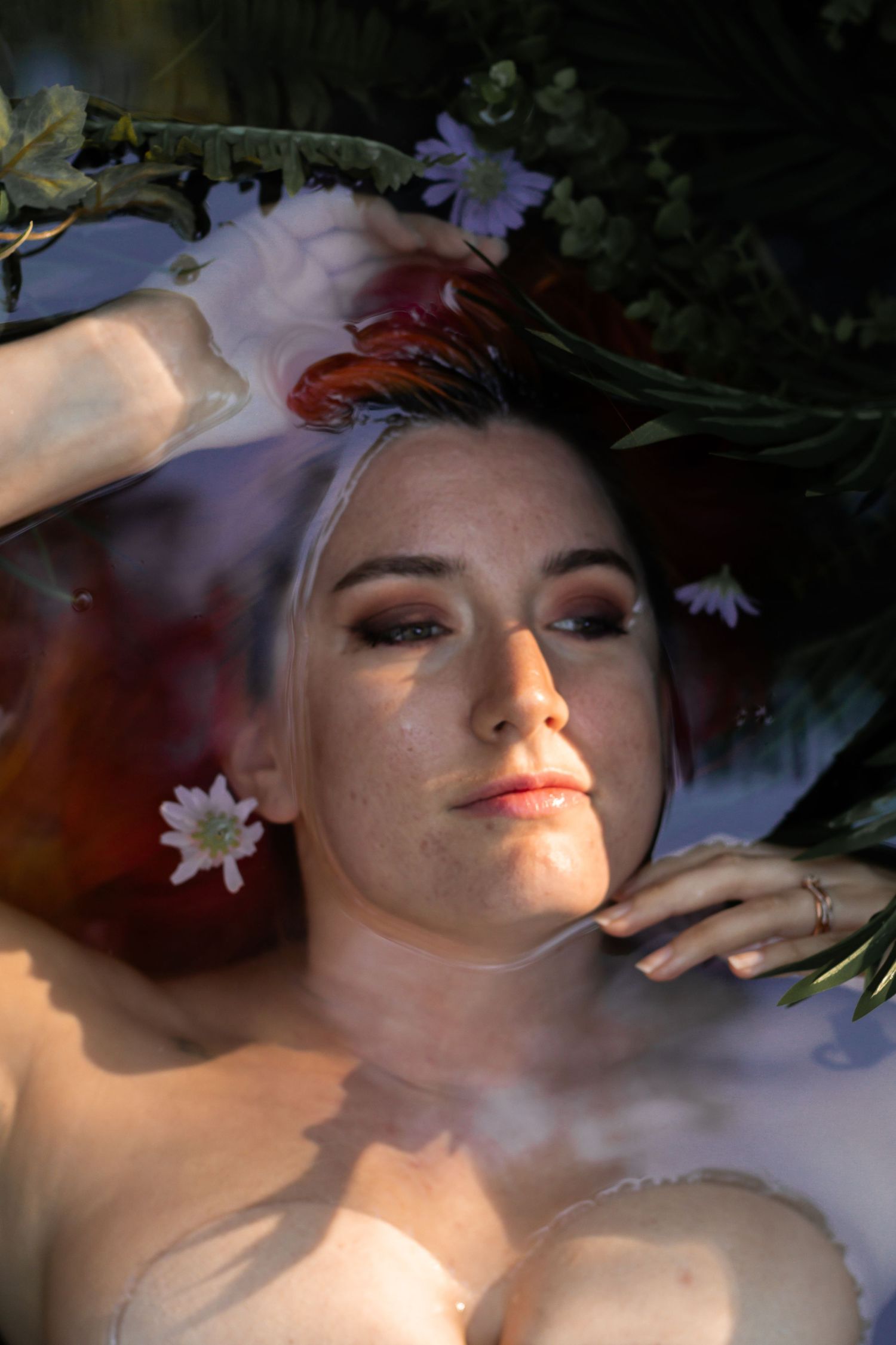 Woman relaxing in a floral milk bath surrounded by green leaves and colorful flowers in soft natural light.
