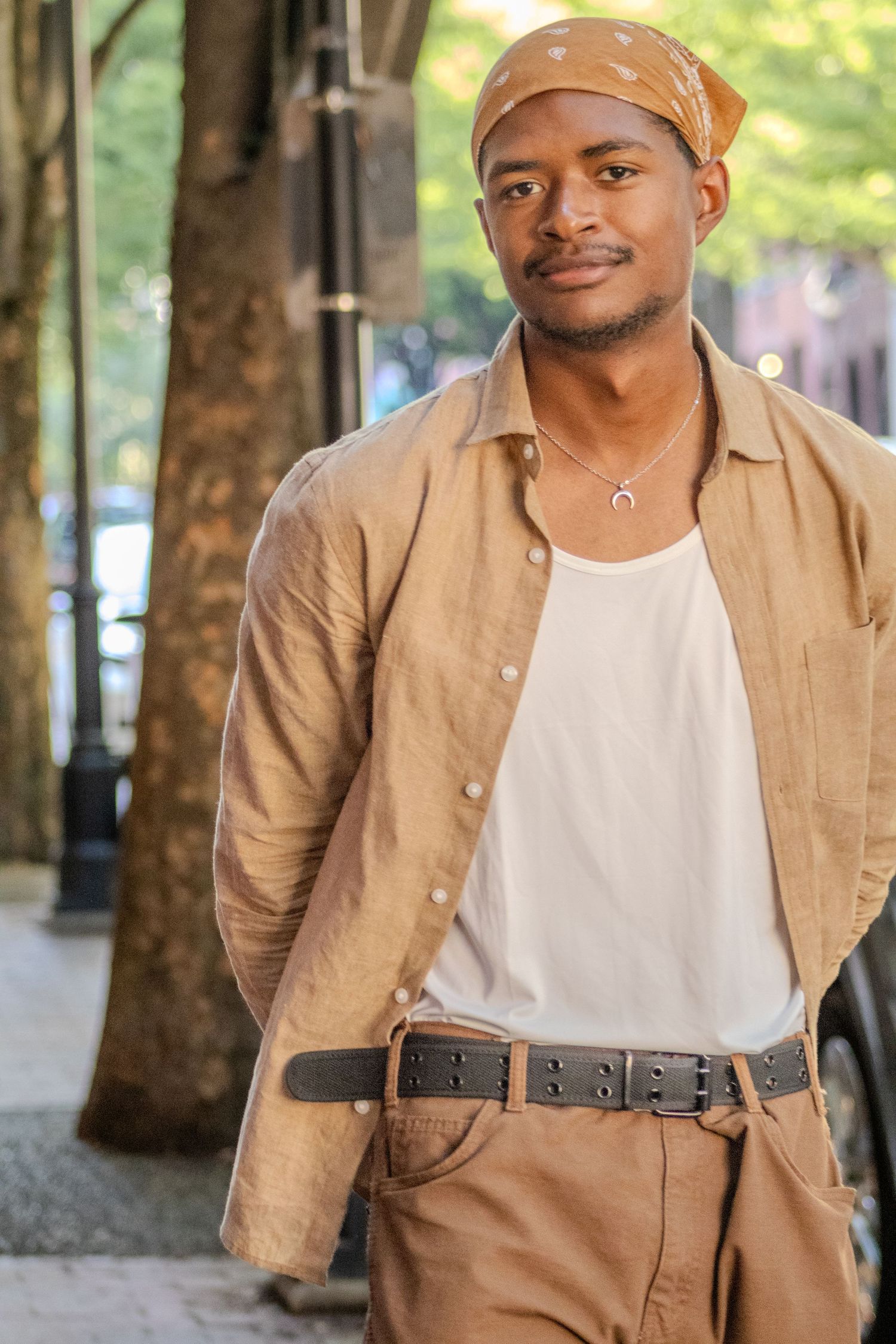 Stylish Black man in tan linen shirt and bandana poses on urban city sidewalk.