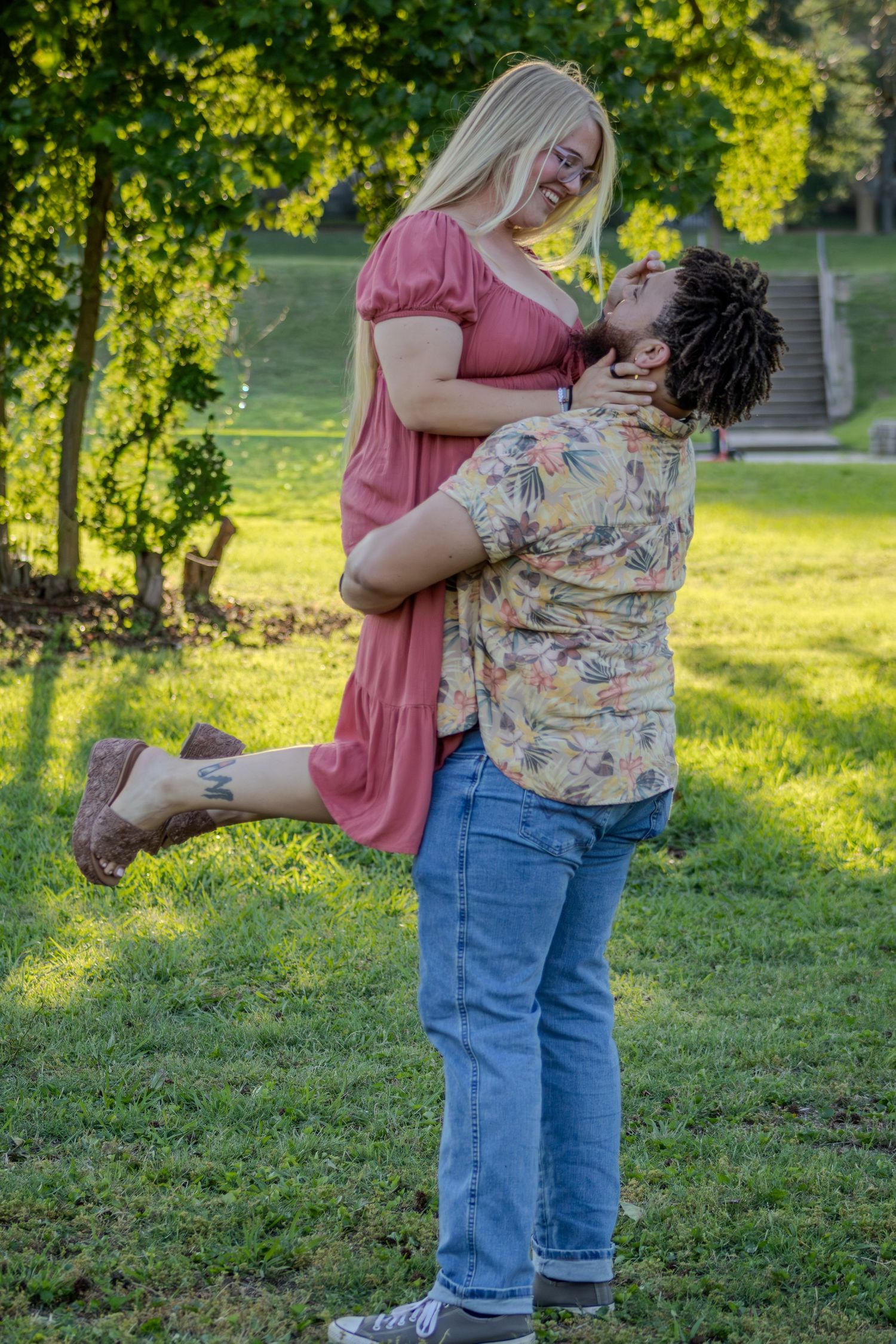 Man lifts smiling blonde woman in pink dress in sunny green park setting.