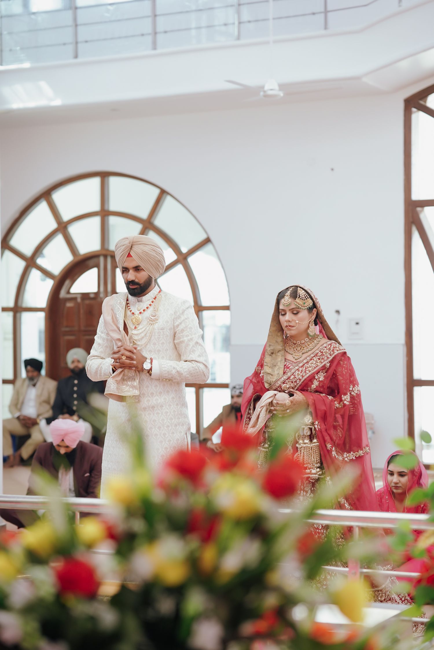 Sikh bride and groom in traditional attire walking during Anand Karaj ceremony in bright white venue.