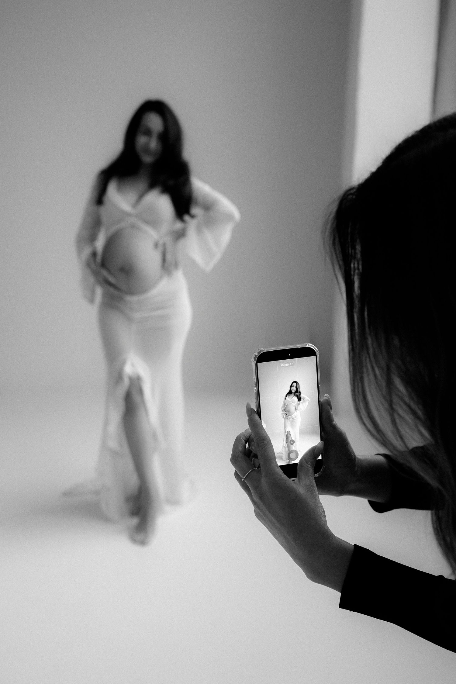 Black and white photo of photographer capturing pregnant woman in white dress on smartphone.