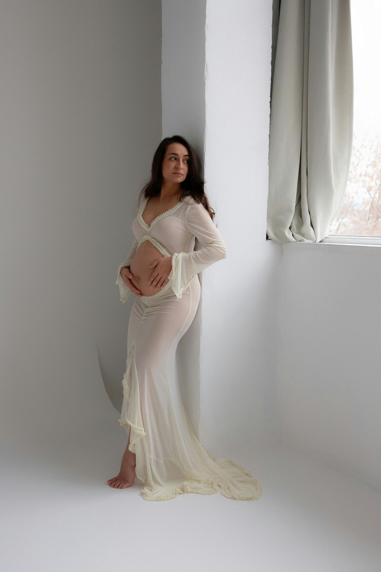 Pregnant woman in sheer white gown standing by window in bright white studio setting.
