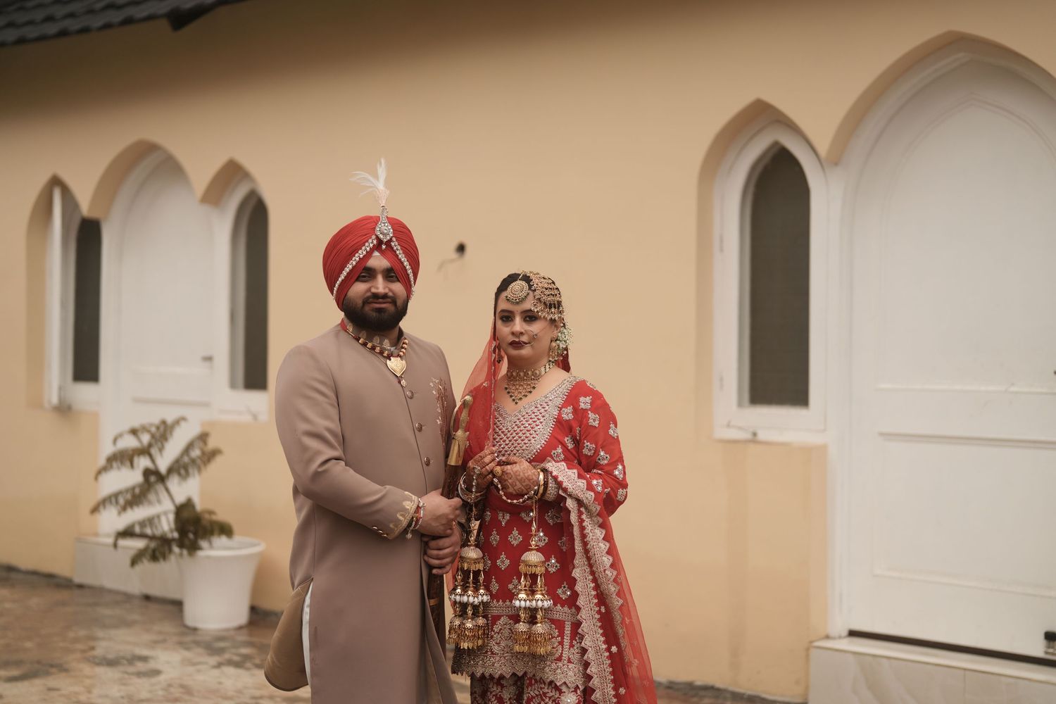 Sikh bride and groom in traditional red and gold wedding attire standing outside a white building.