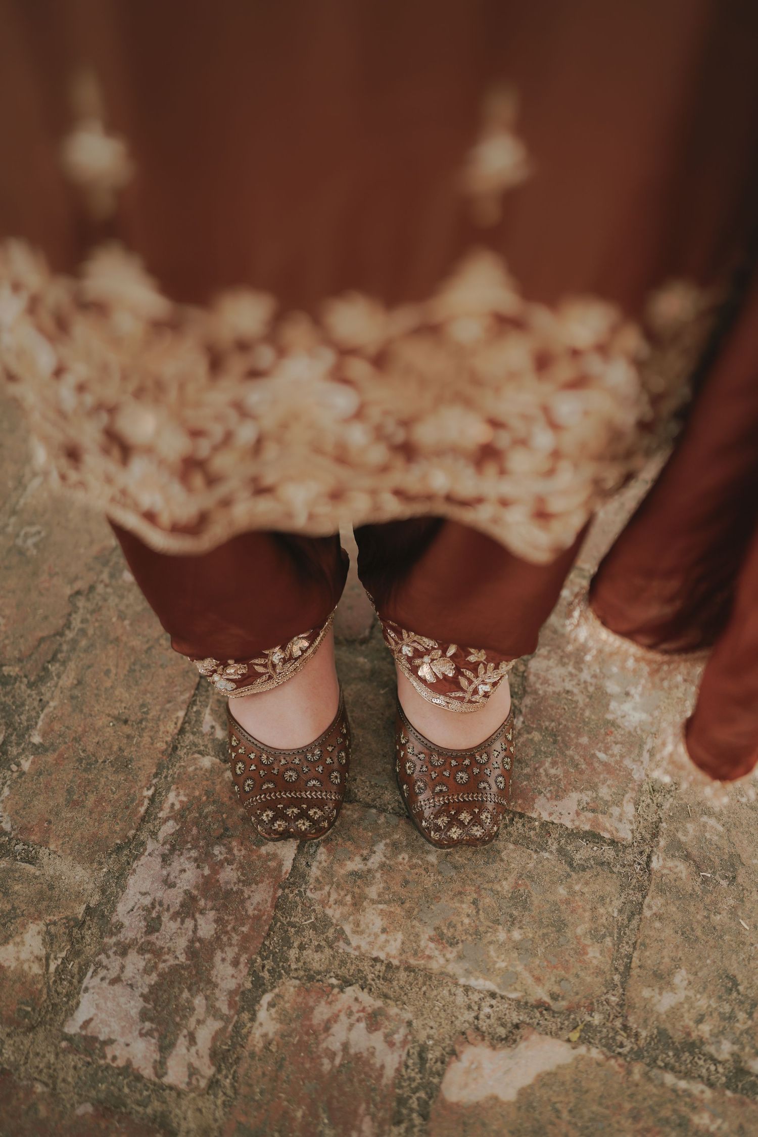 Close-up of bride's embroidered brown juttis on cobblestone floor with lace kurta hem.
