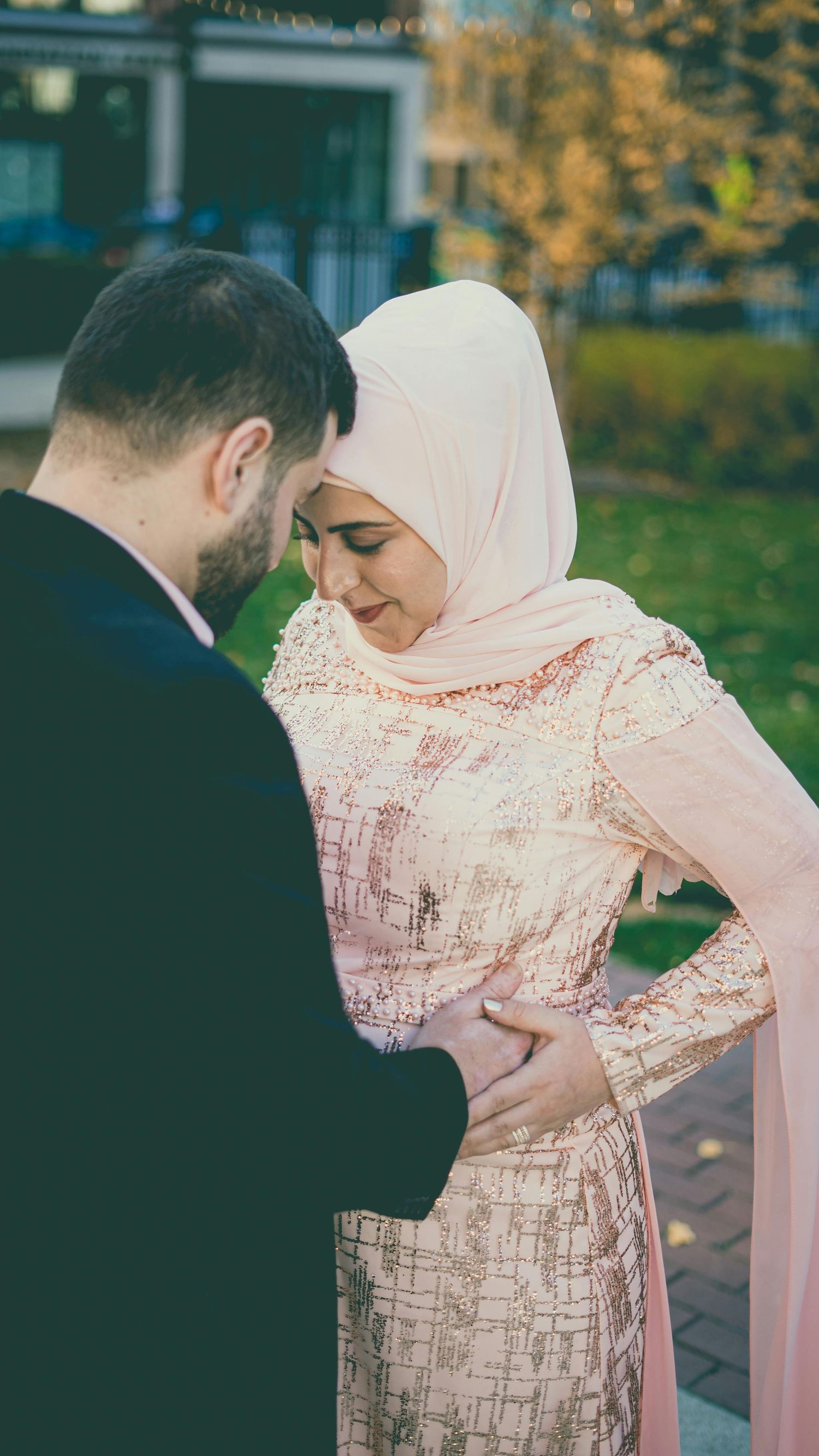 Muslim couple sharing tender moment outdoors, woman in pink hijab and sequined dress smiling.