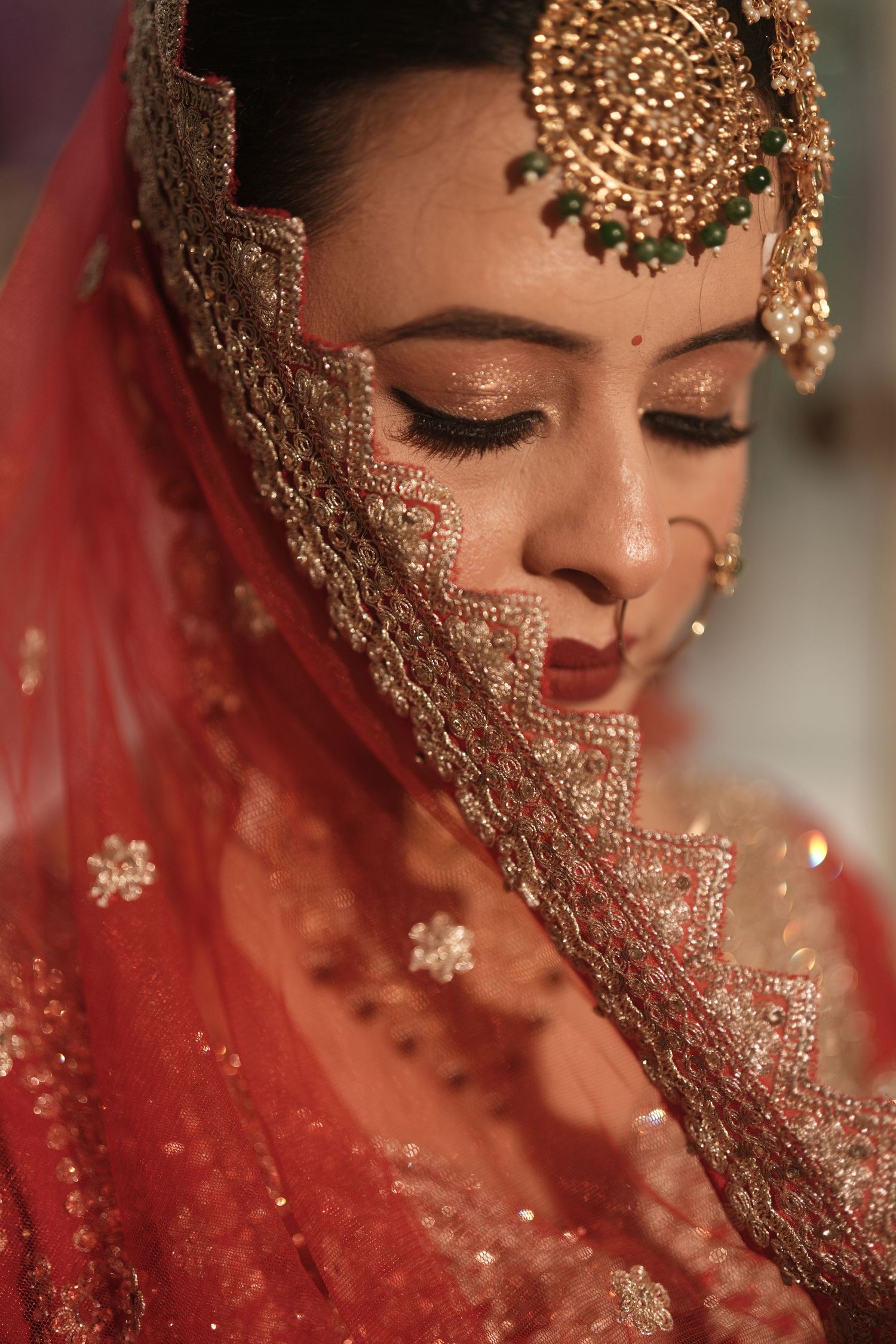 Indian bride in red dupatta with gold maang tikka and traditional bridal jewelry looking down.