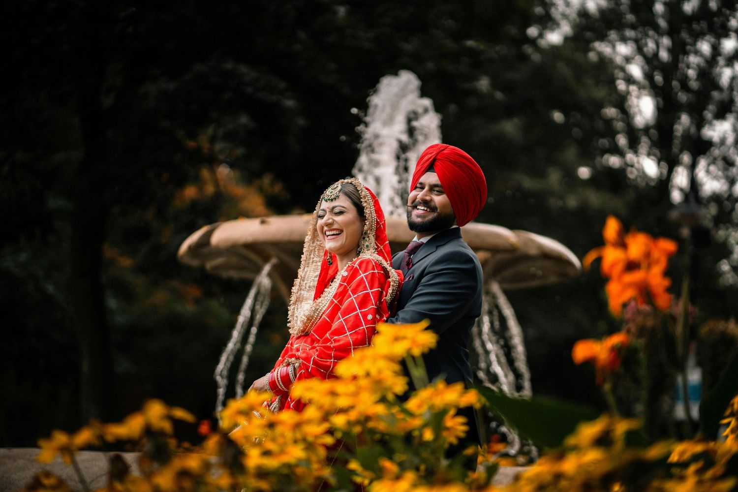 Sikh bride in red lehenga and groom in red turban posing joyfully near fountain with autumn flowers.