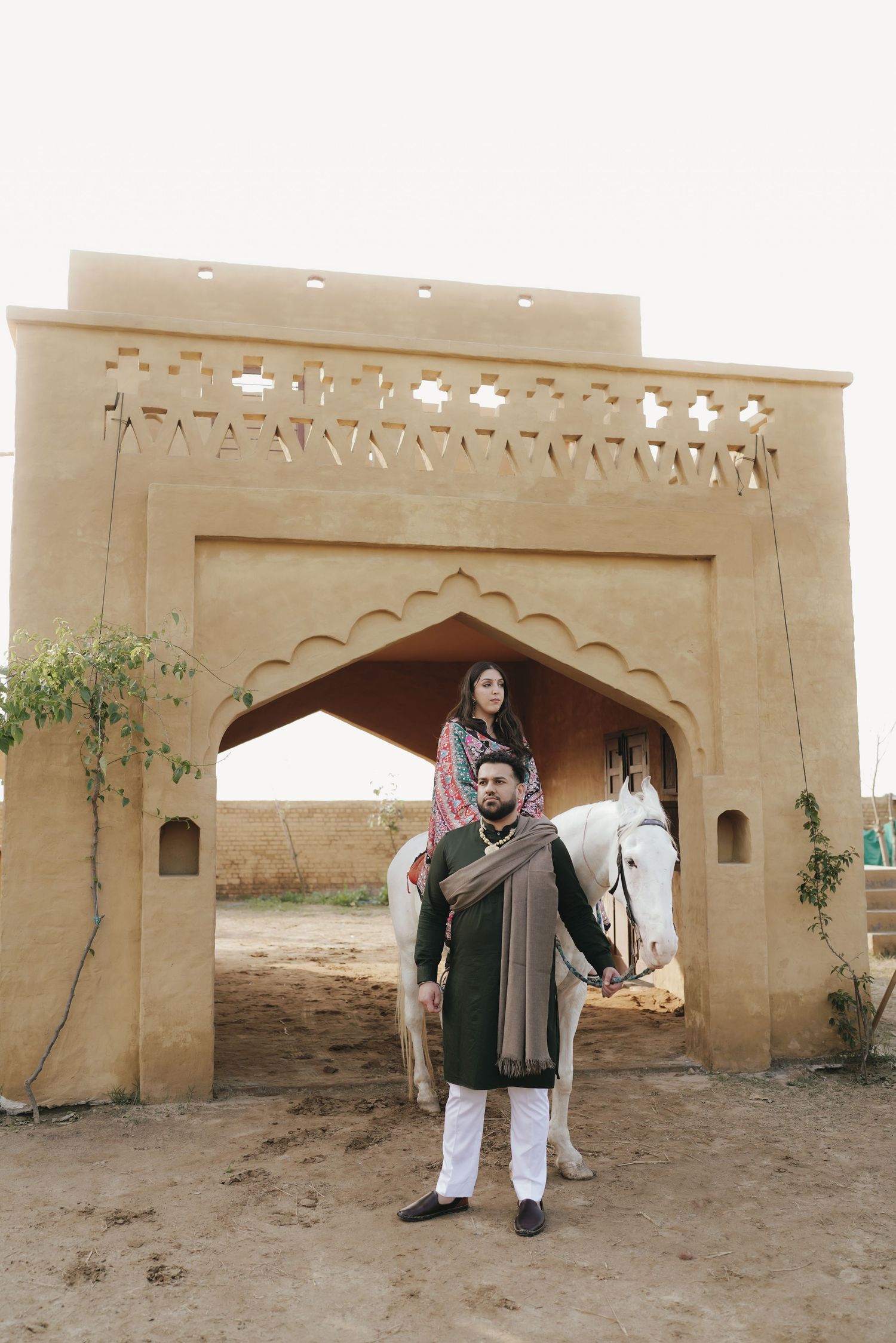 South Asian couple posing at an ornate sandstone archway with a white horse during a pre-wedding photoshoot.