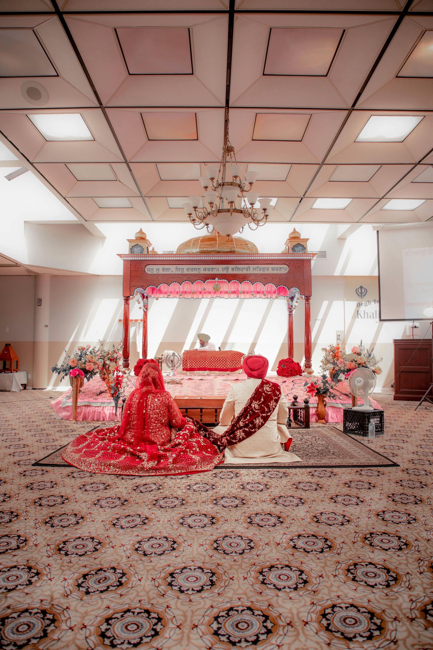 Sikh bride and groom seated before the Guru Granth Sahib during Anand Karaj ceremony indoors.