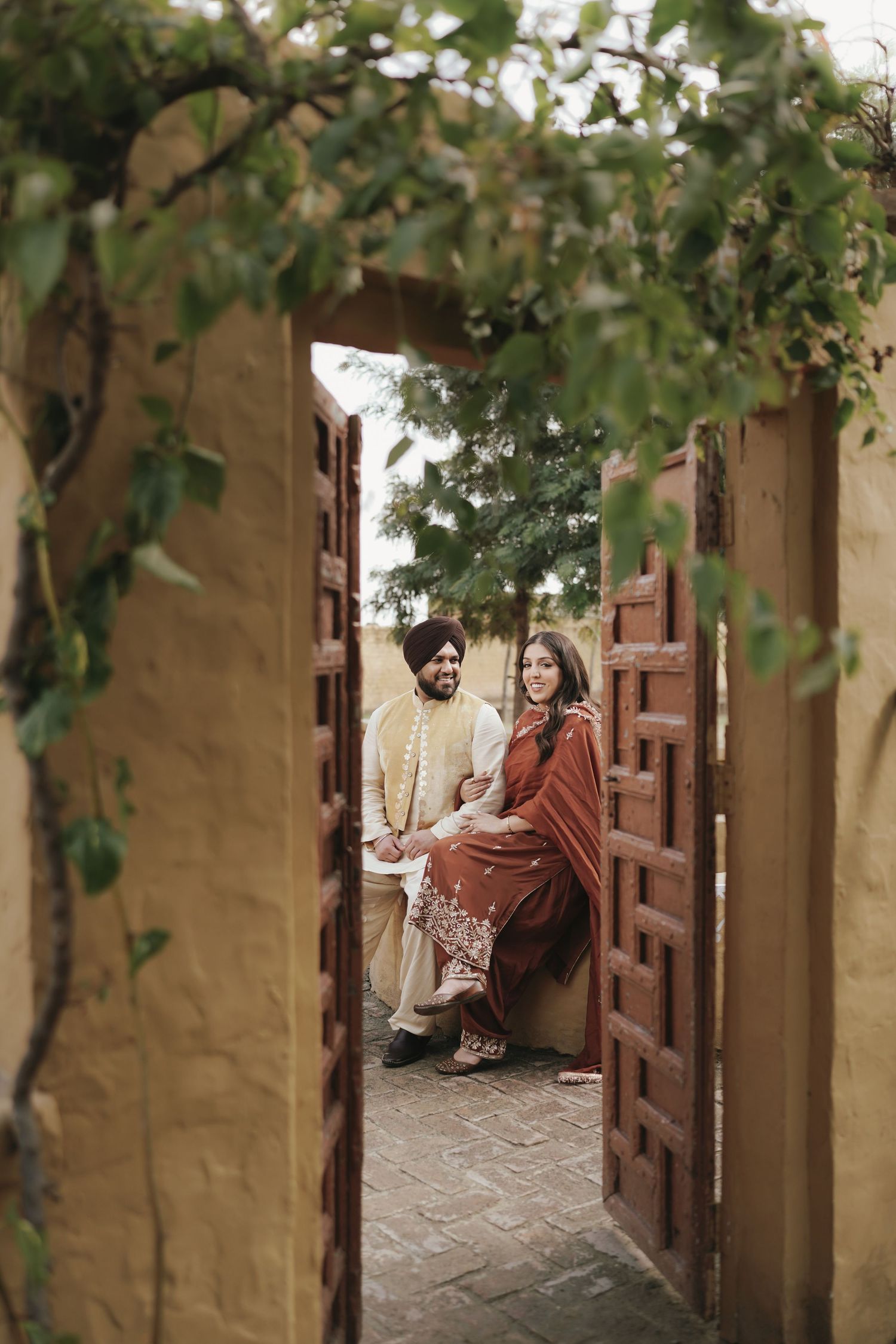Sikh couple in traditional attire sitting together framed by wooden doors and green foliage.