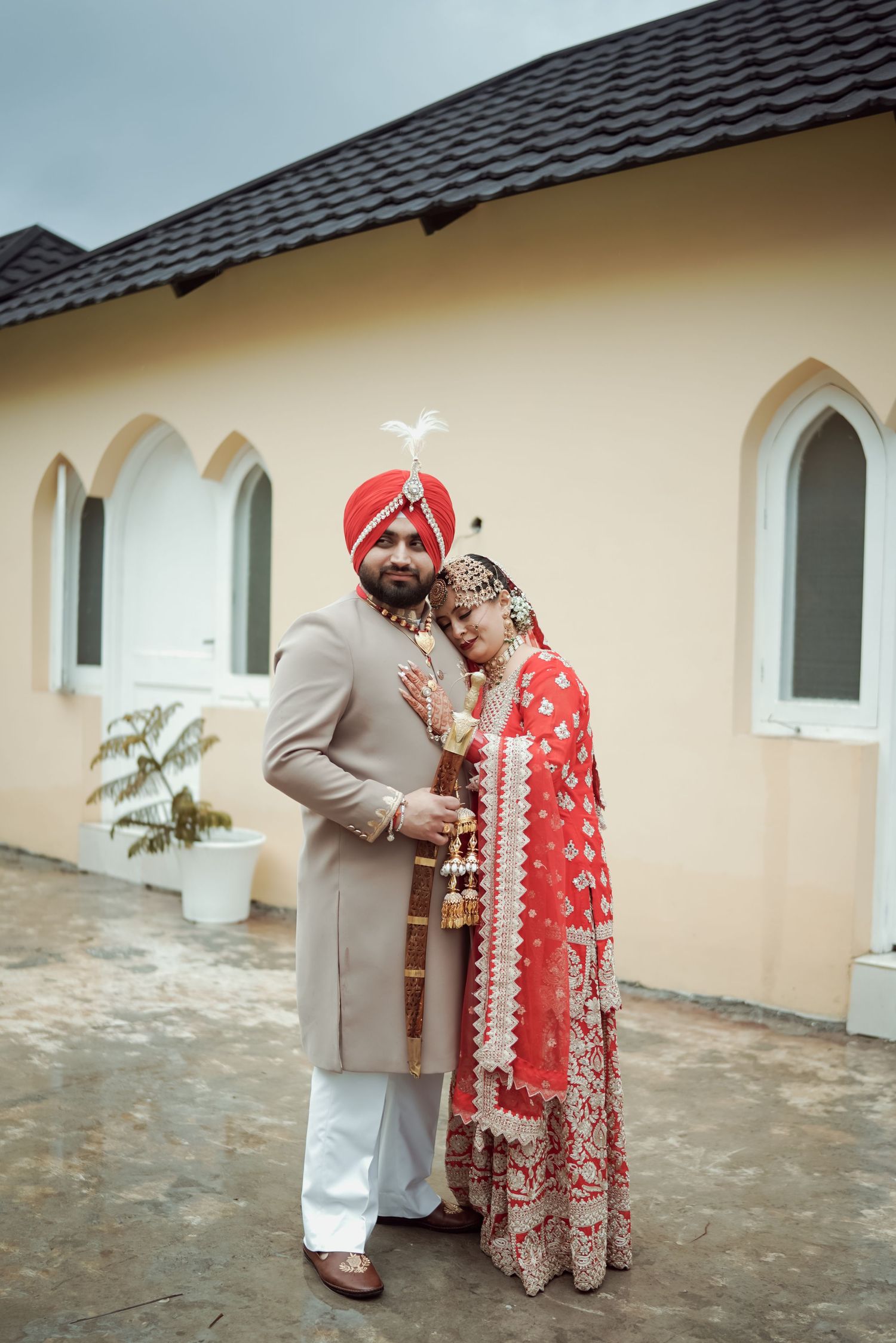 Sikh wedding couple in traditional attire, bride in red lehenga leaning on groom outdoors.