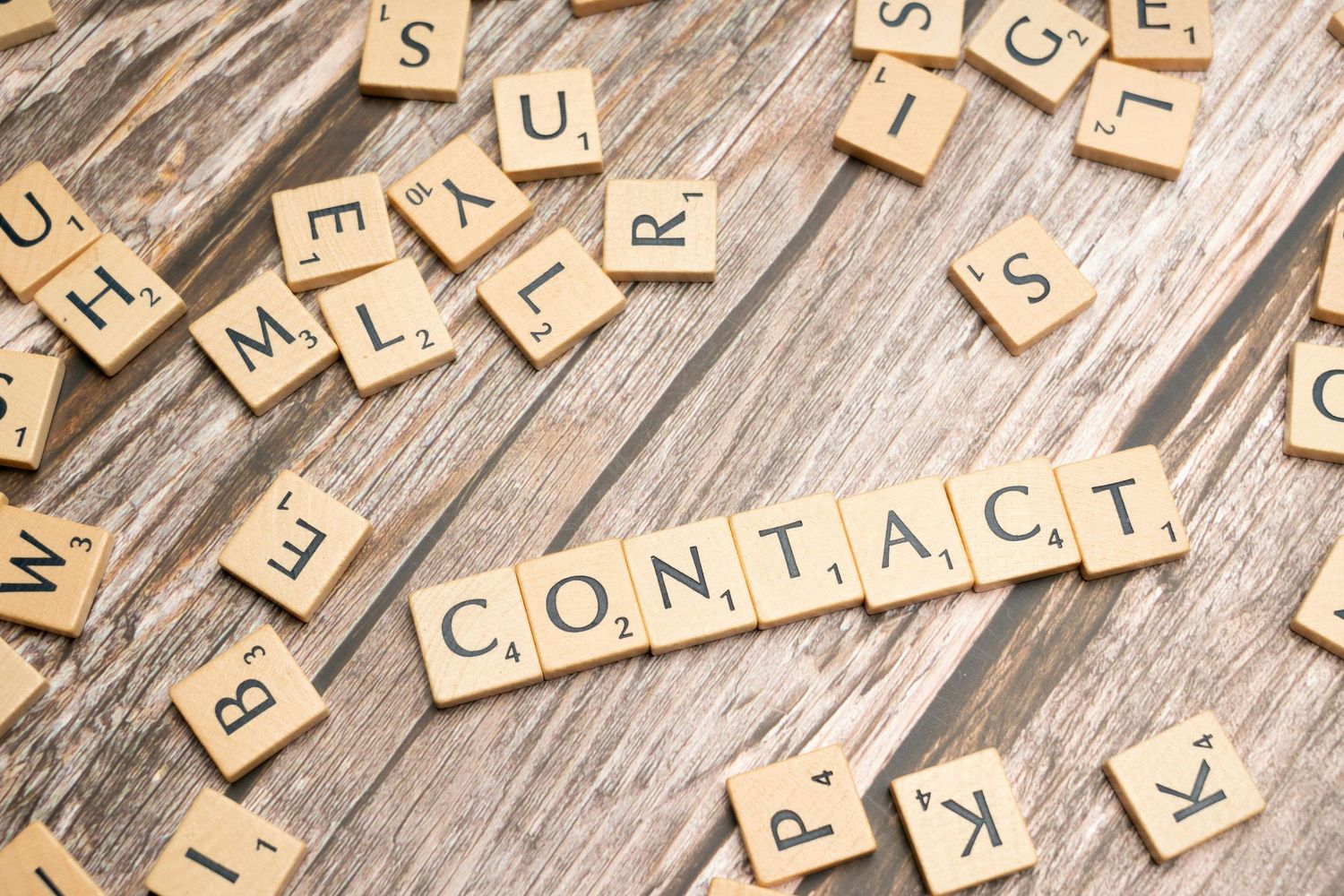 Scrabble tiles spelling CONTACT on rustic wooden surface with scattered letter tiles.