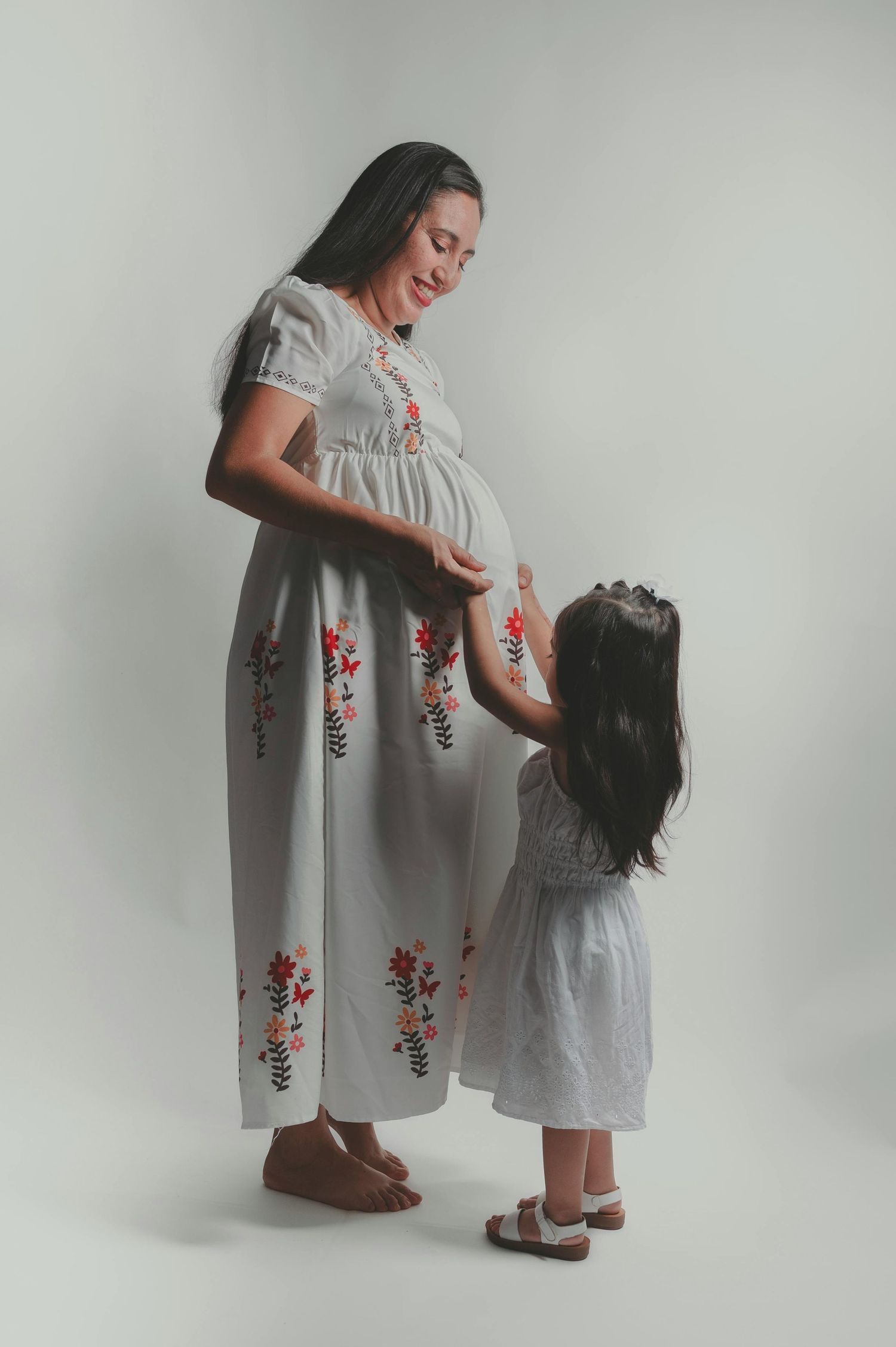 Pregnant woman in floral white dress smiling at young daughter touching her baby bump.