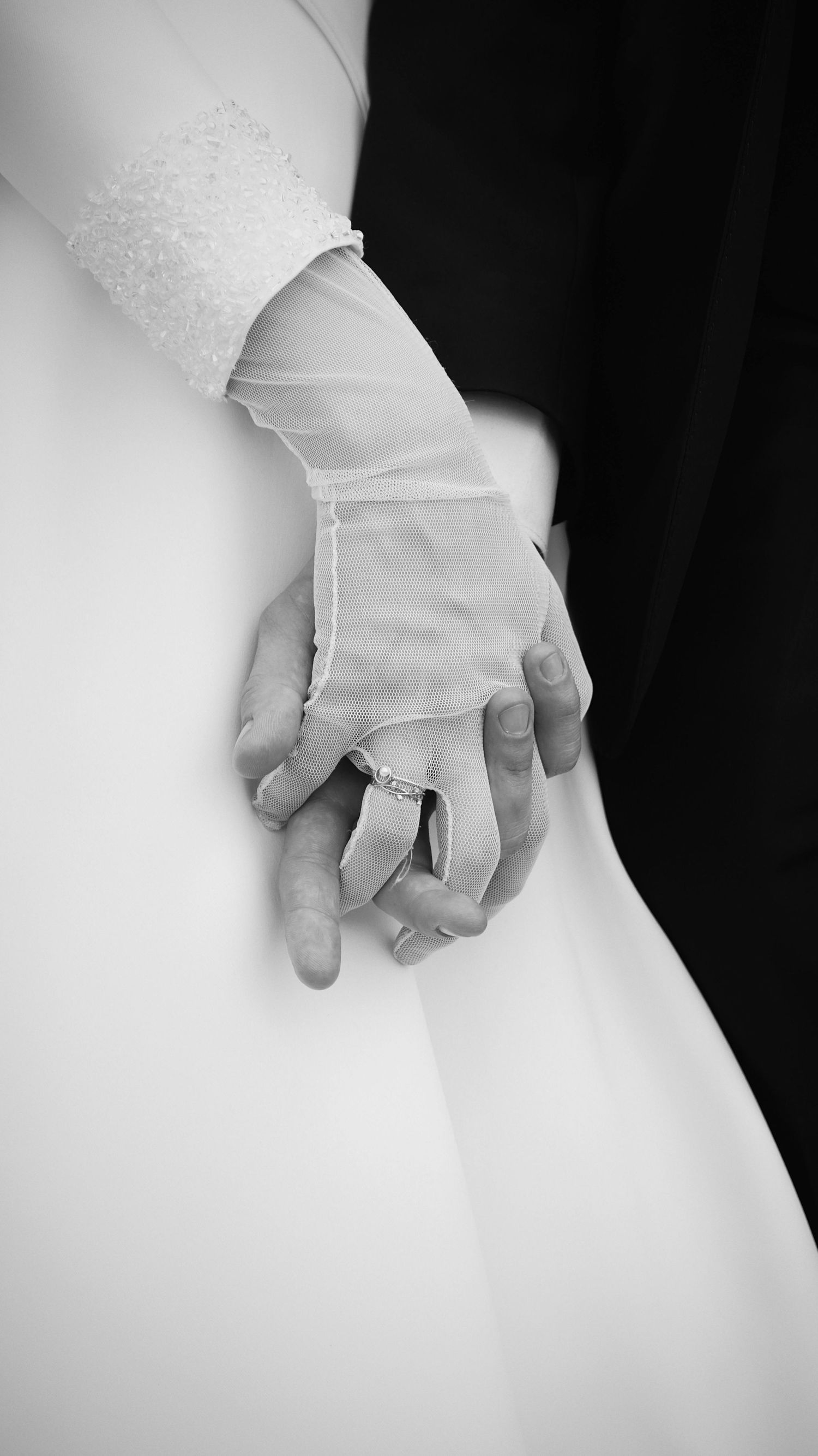 Black and white close-up of bride and groom holding hands with lace glove and diamond ring.