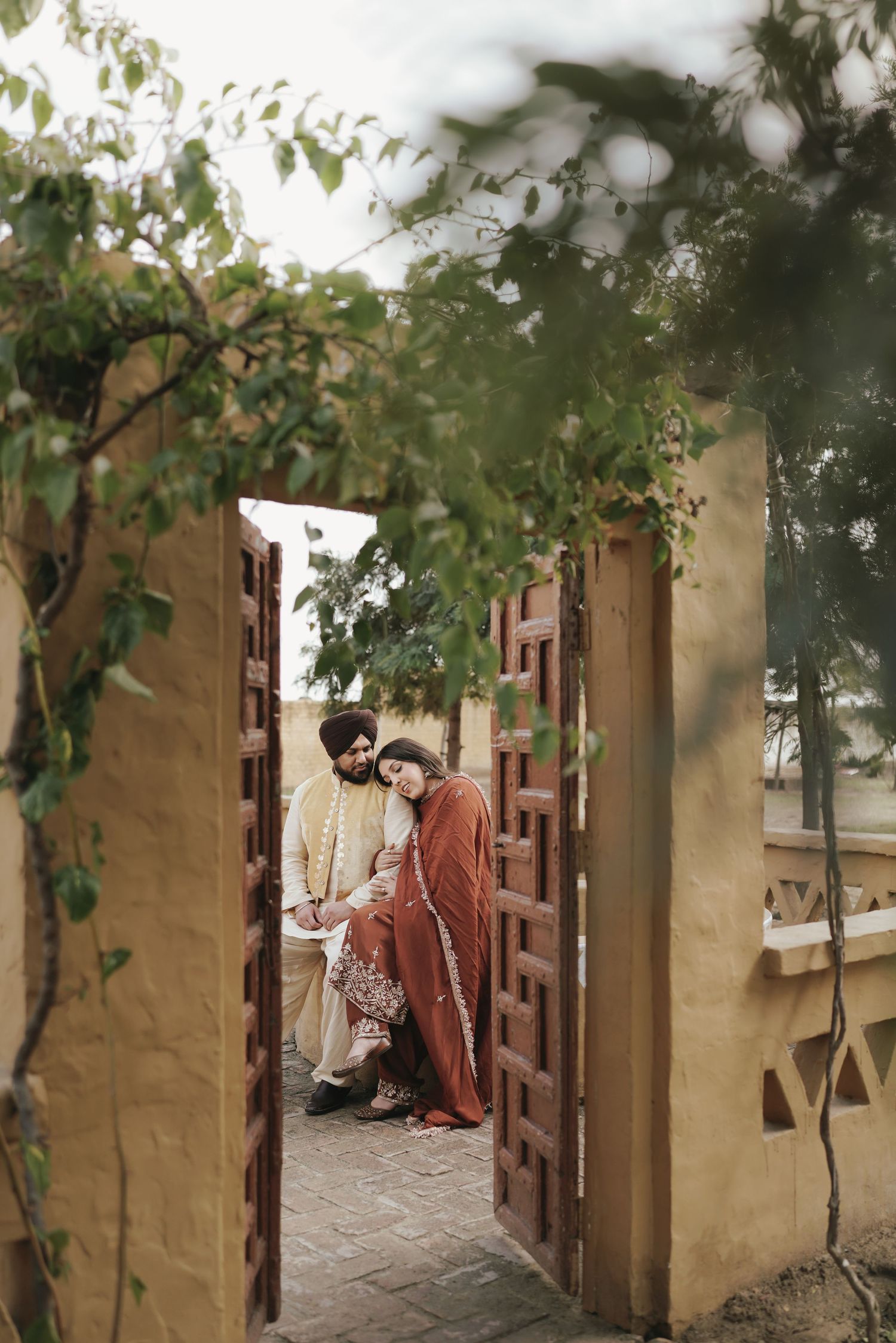 Couple in traditional Indian attire sharing a tender moment framed by an ornate wooden doorway with lush greenery.