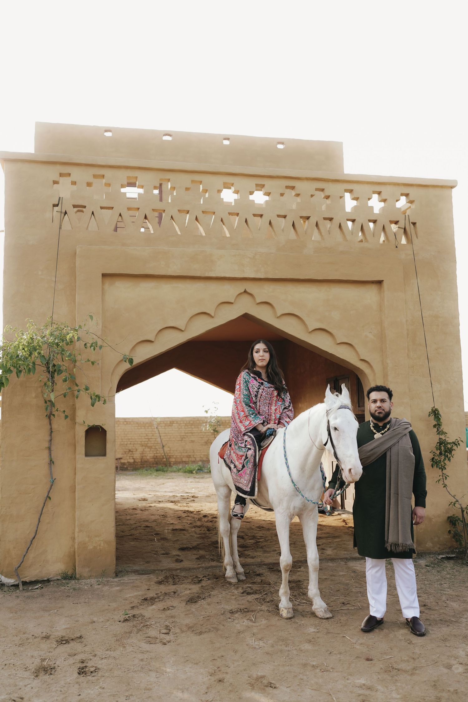 Woman in embroidered suit riding white horse under arched gateway with man standing beside.