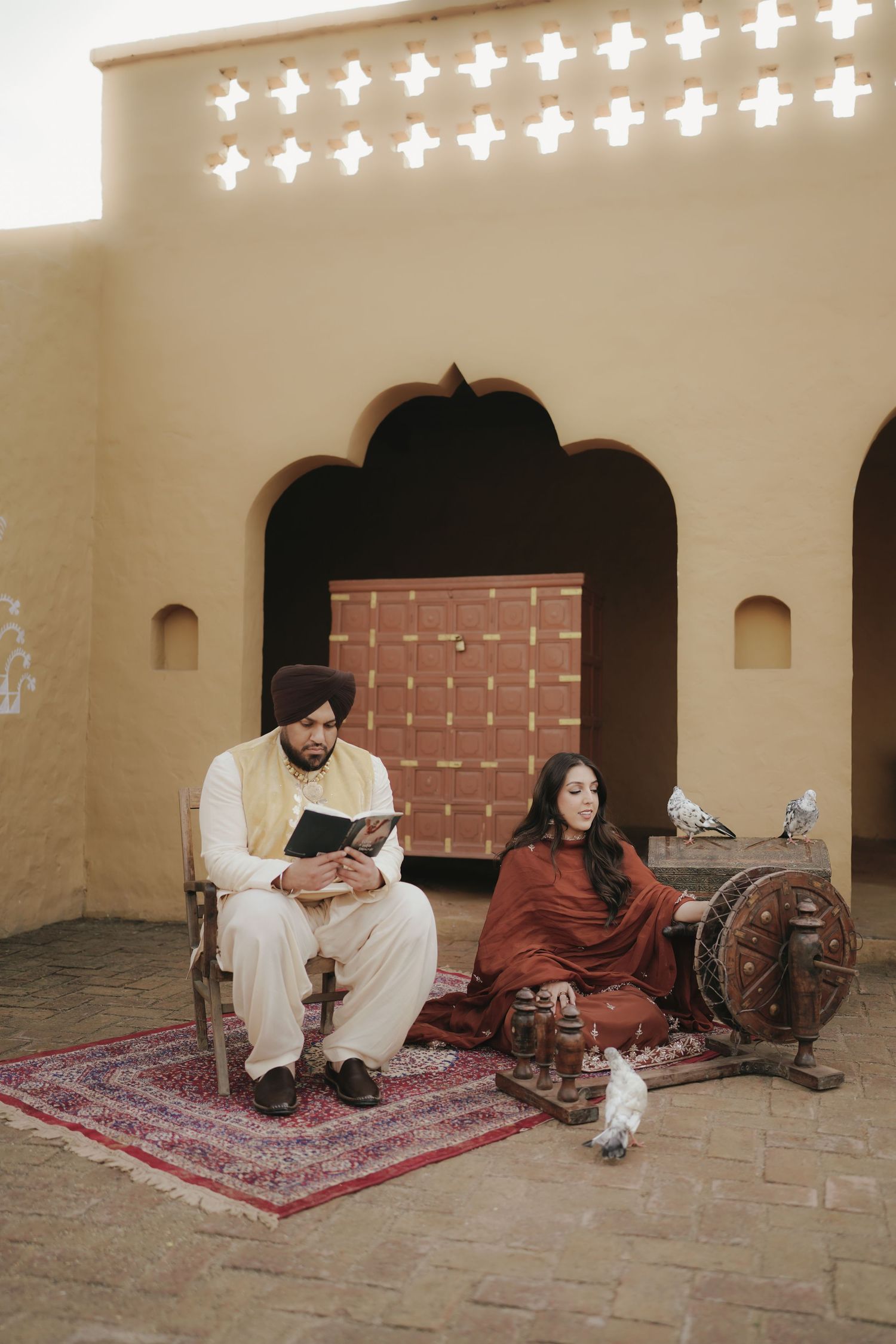 Sikh couple in traditional attire sitting in courtyard, man reading book, woman at spinning wheel.