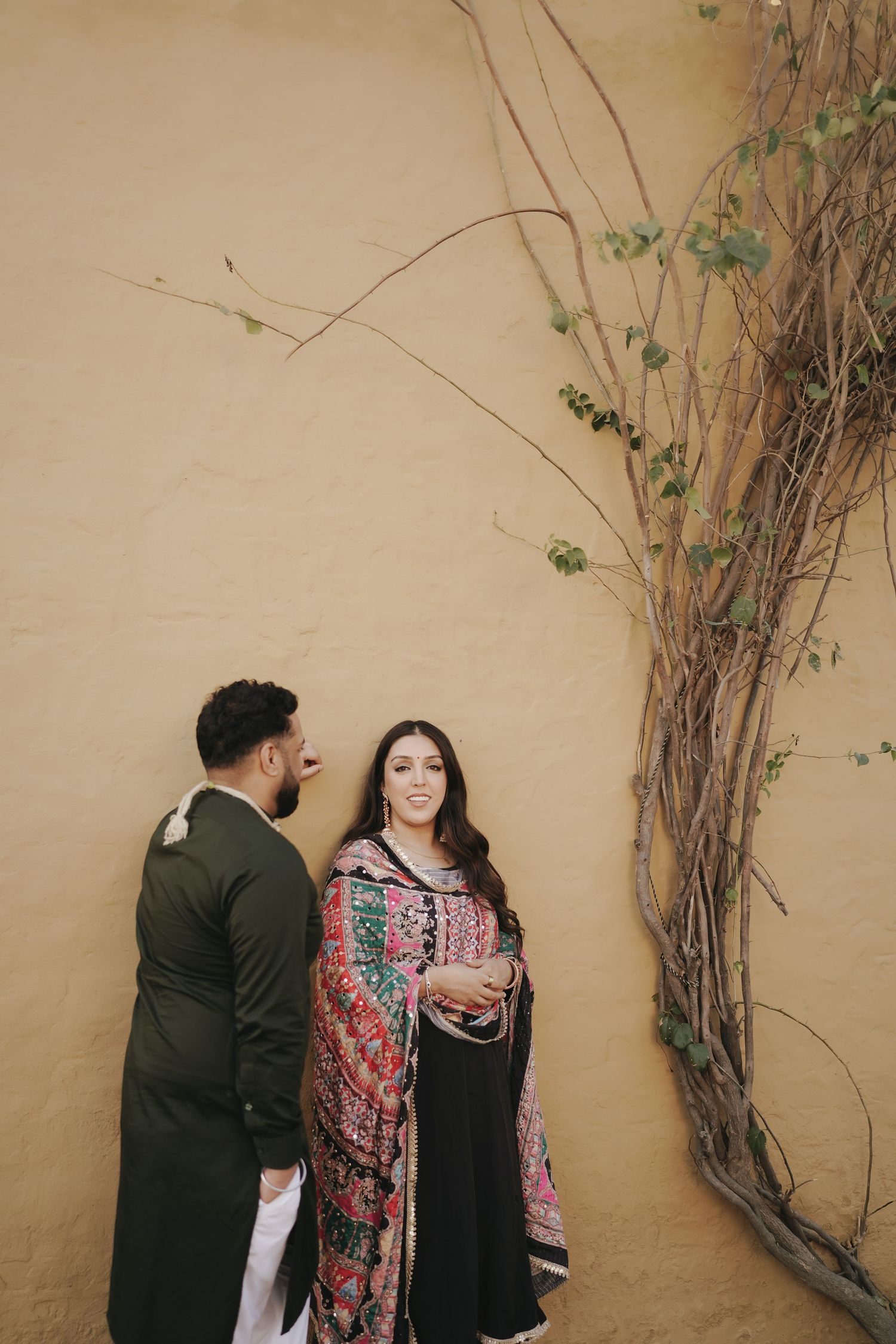 South Asian couple in traditional attire standing by a warm yellow wall with climbing vines during a pre-wedding shoot.