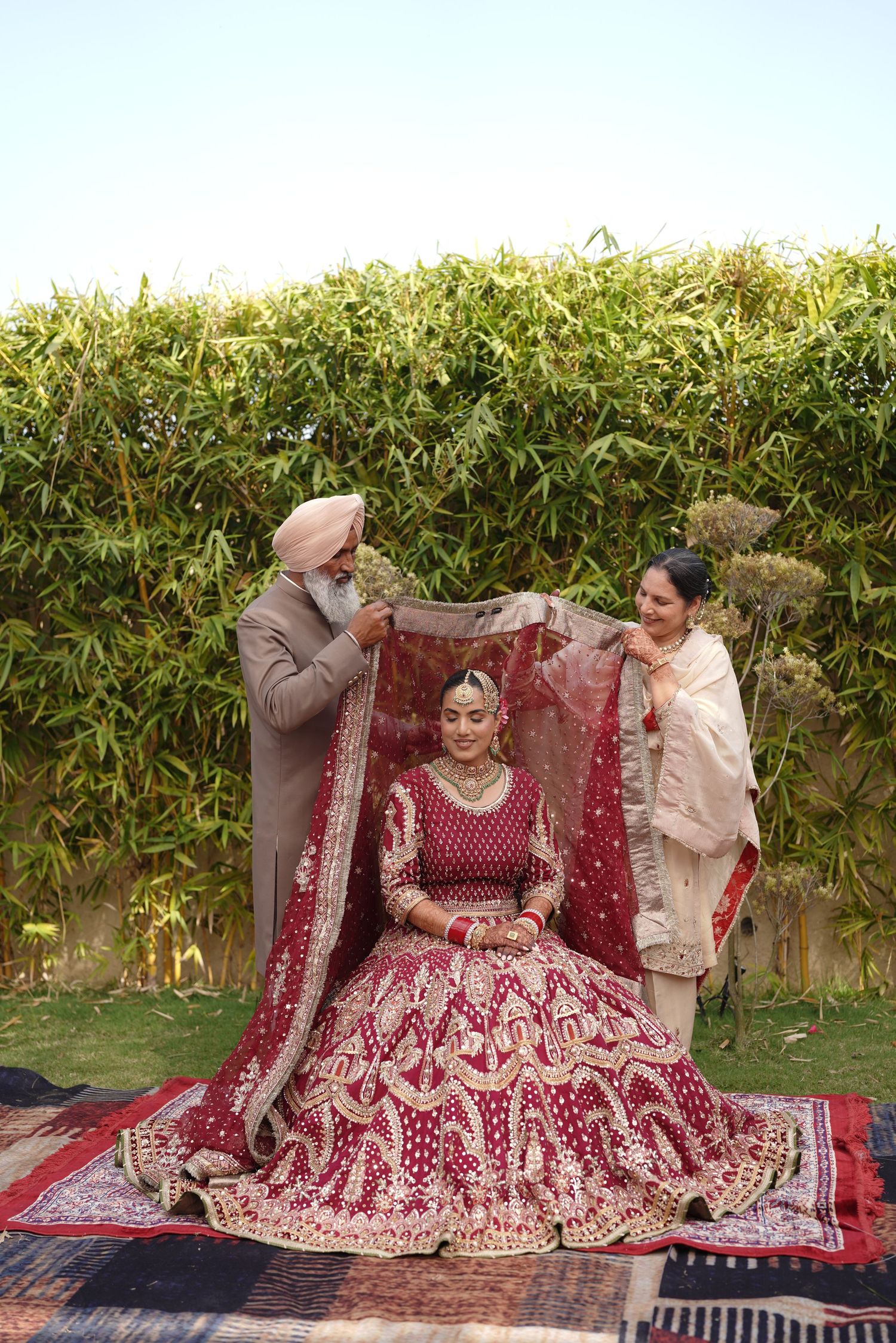 Sikh bride in red lehenga seated while parents drape dupatta in garden setting.